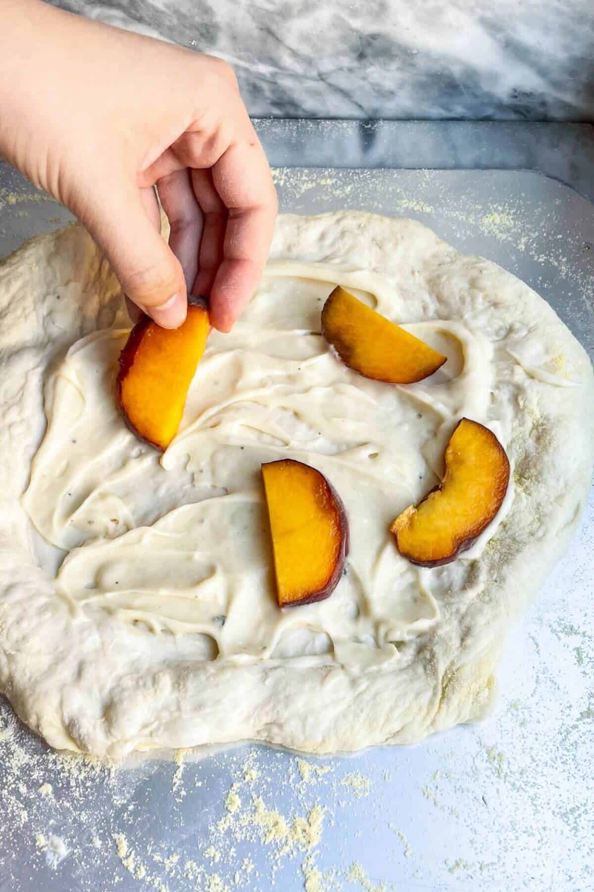 Hands placing peach slices onto pizza base sitting on a steel pizza peel.