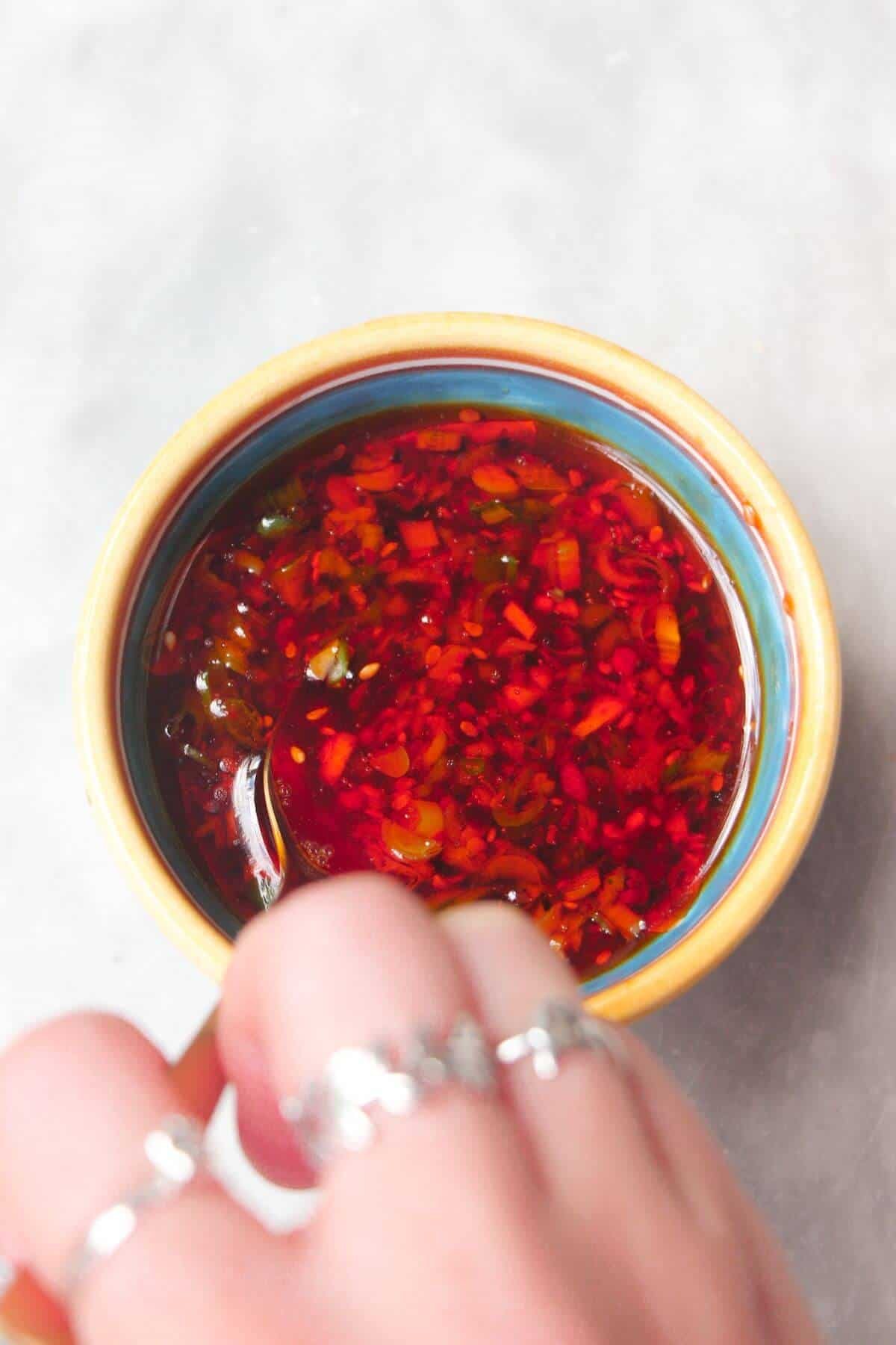 Hand holding a spoon, mixing red dumpling dipping sauce in a small bowl.