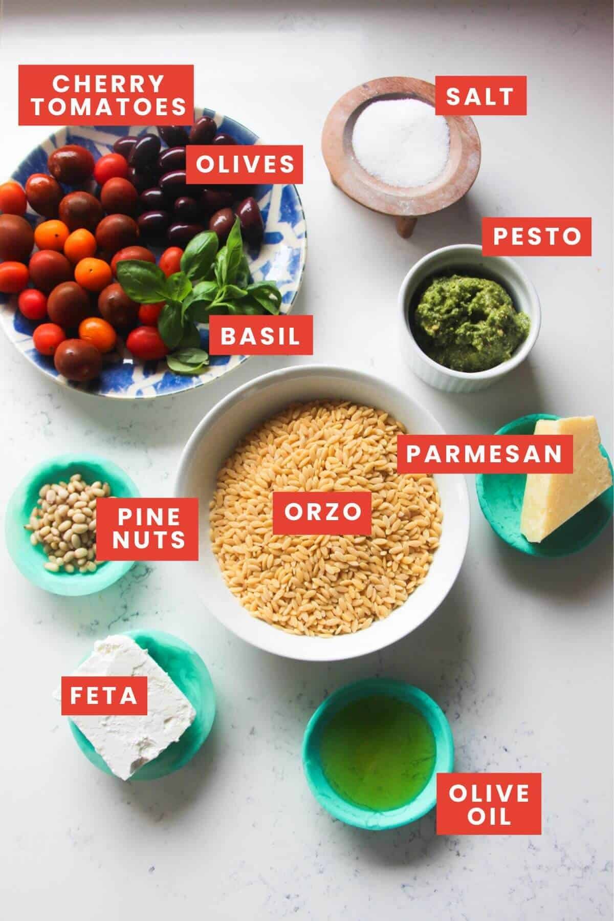 Ingredients for orzo pesto salad laid out on a white marble background and labelled.
