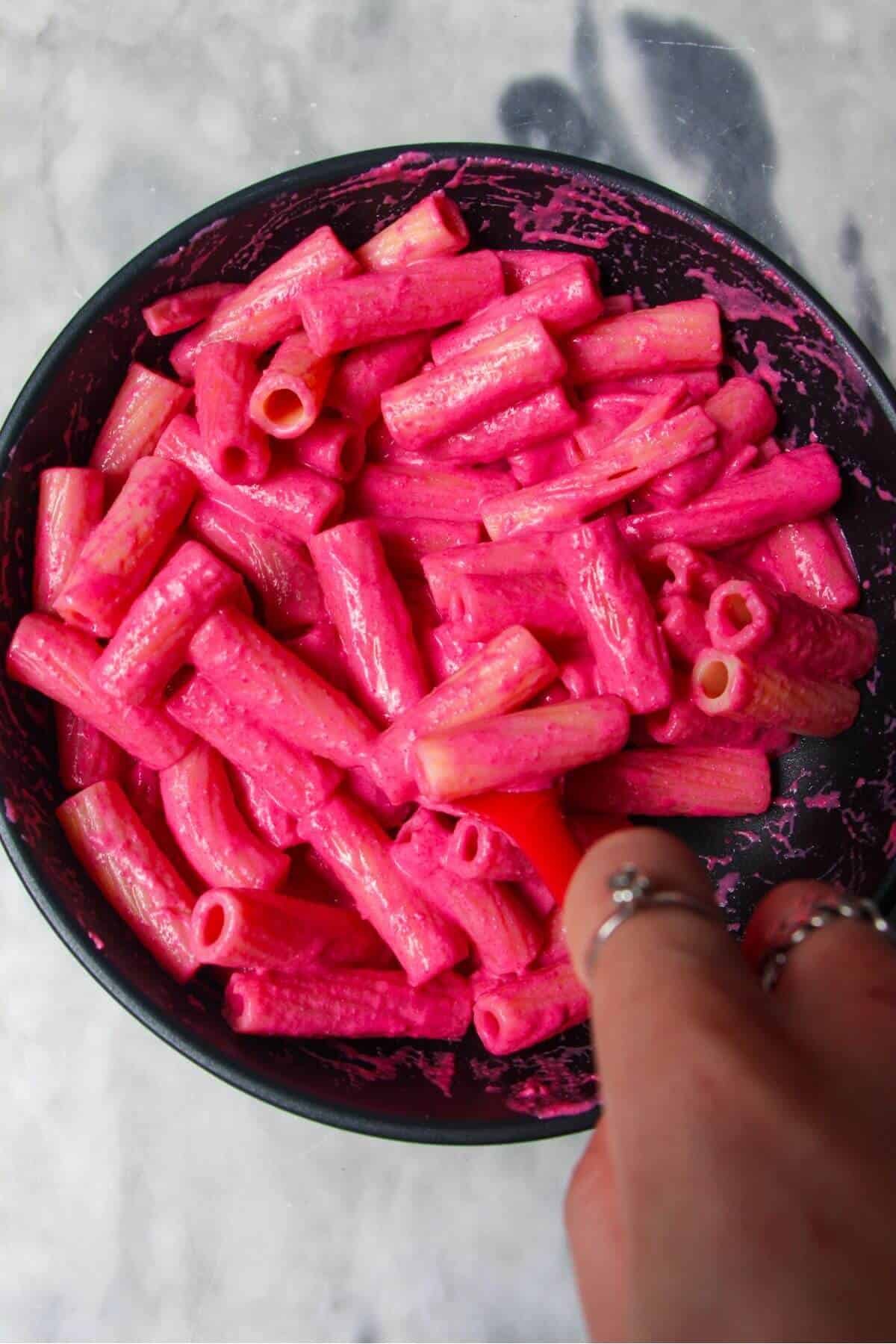 Hand holding red spatula stirring pink sauce into pasta in a small black pan.