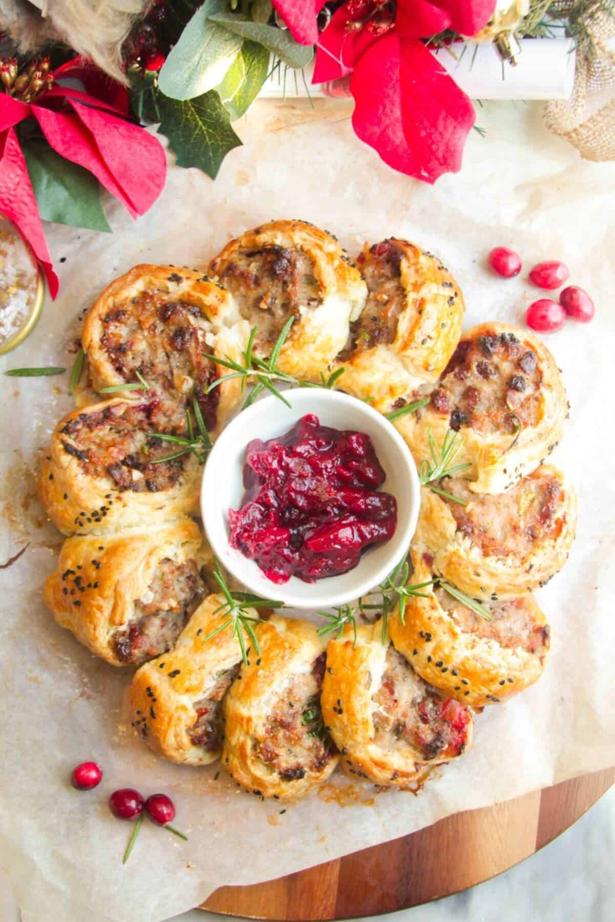 Christmas sausage roll wreath on baking paper on a wooden board with Christmas decorations in the backgorund.