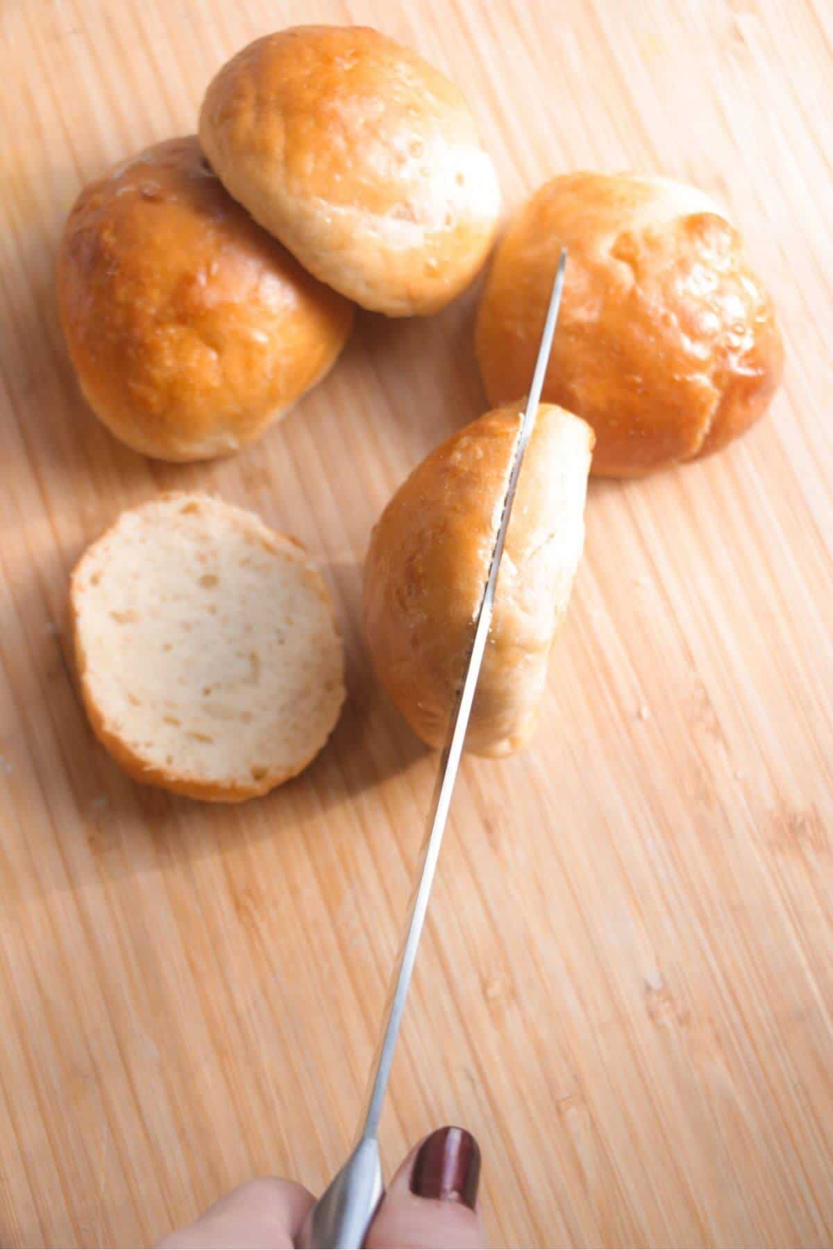 Slider buns on a wooden board, being sliced with a bread knife.