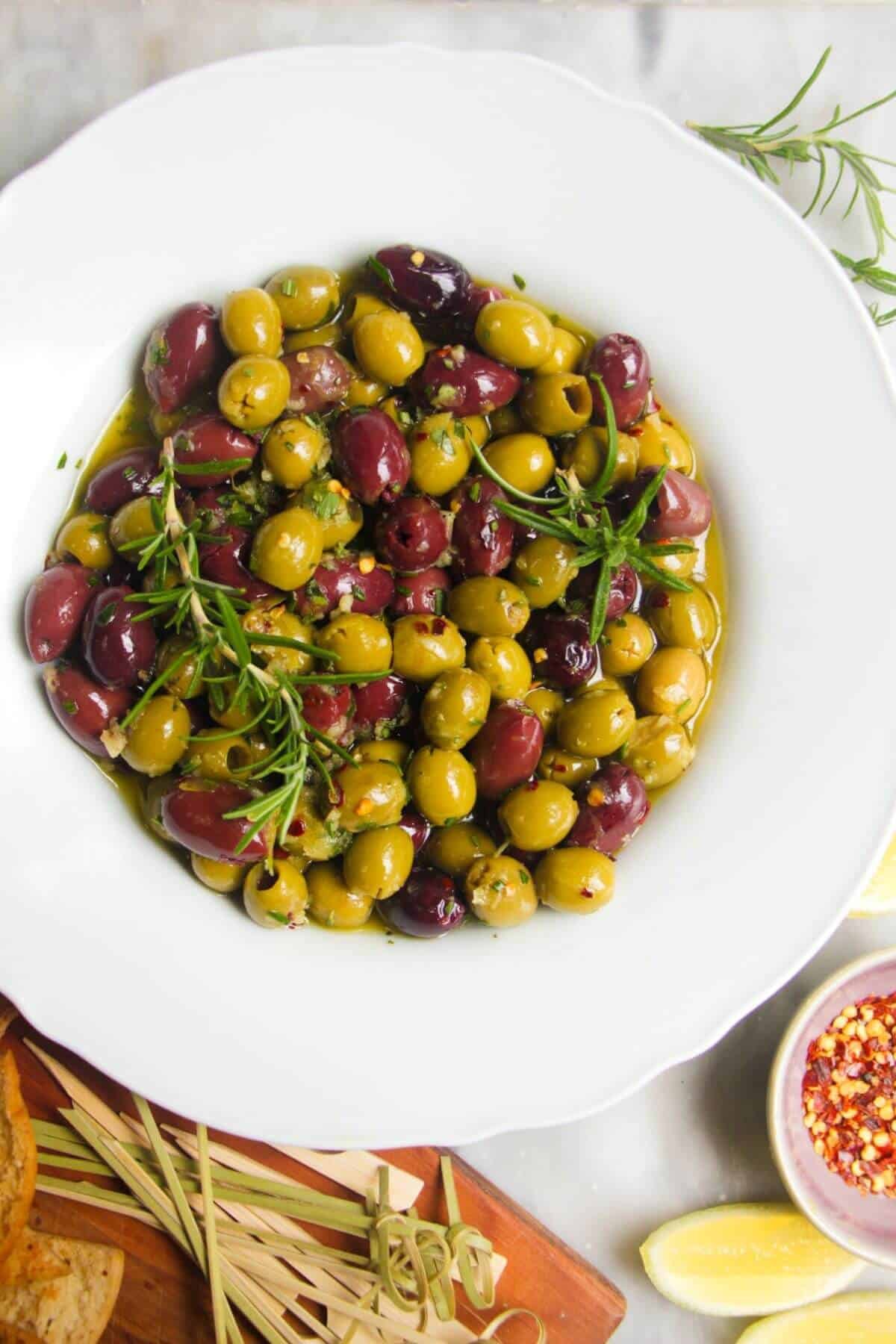 Close up of a white scalloped bowl of marinated olives, with skewers on the side.