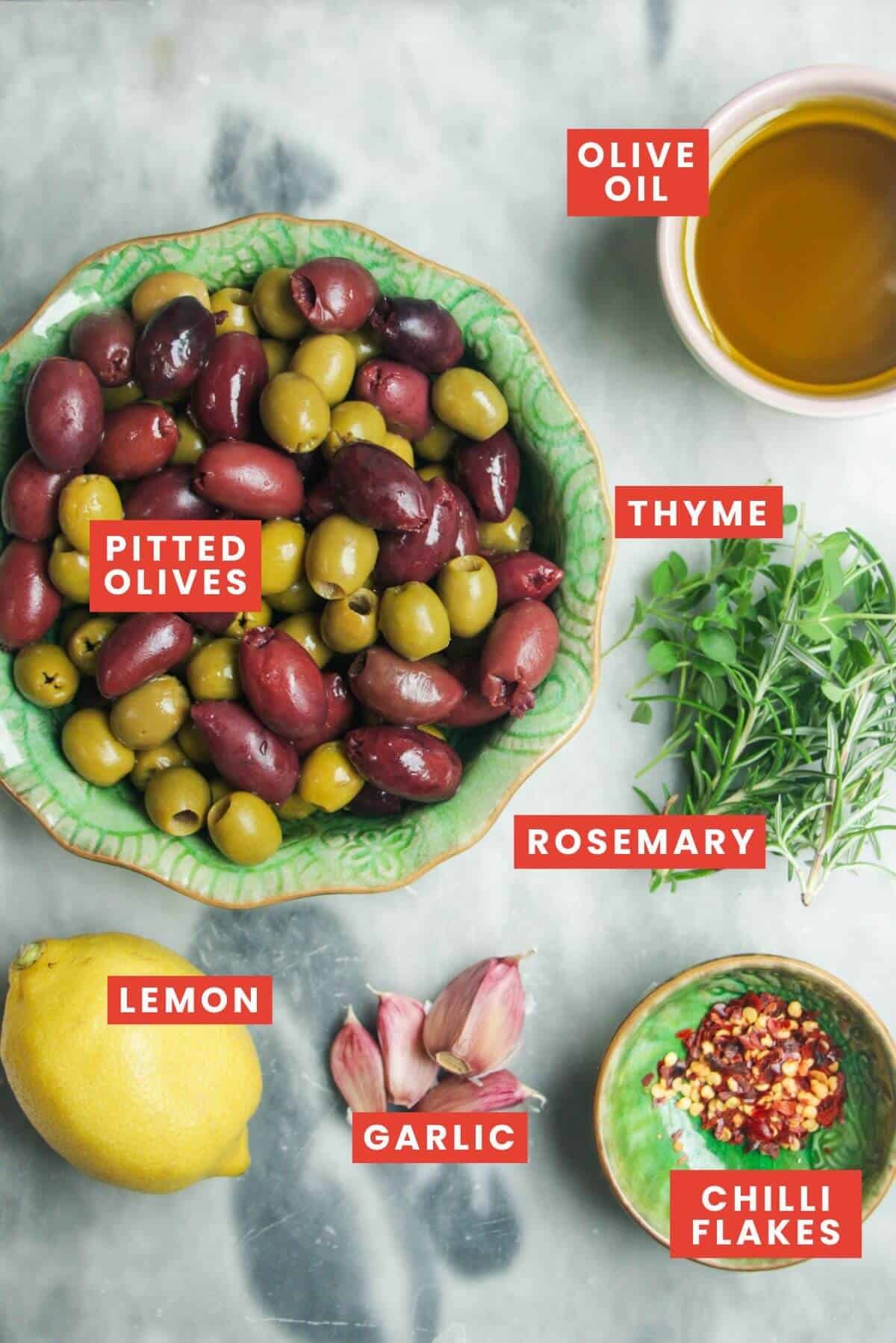 Ingredients for marinated olives on a grey marble background and labelled.