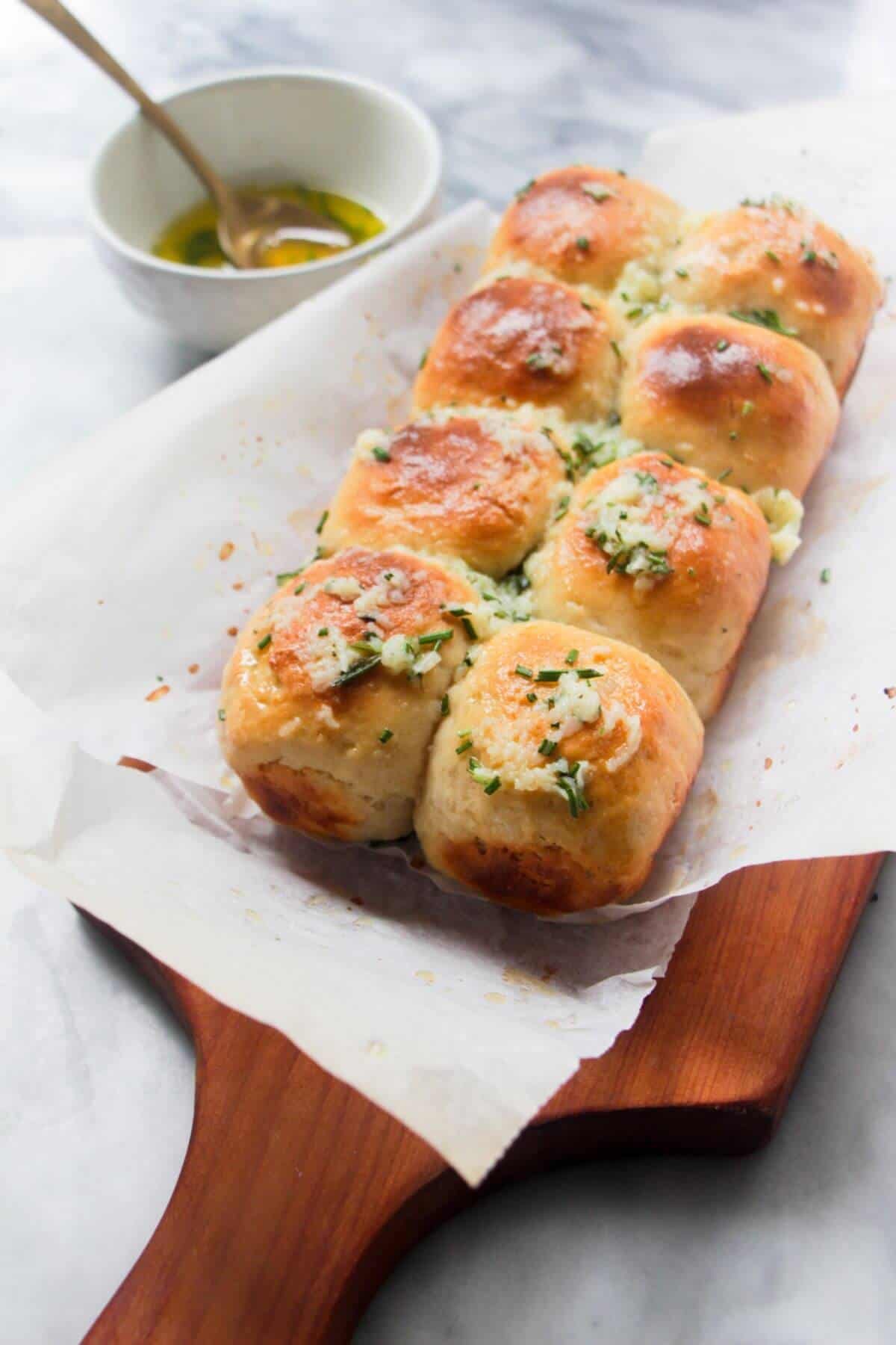 8 small rolls topped with garlic butter on a baking paper lined wooden board with bowl of garlic butter in the background.