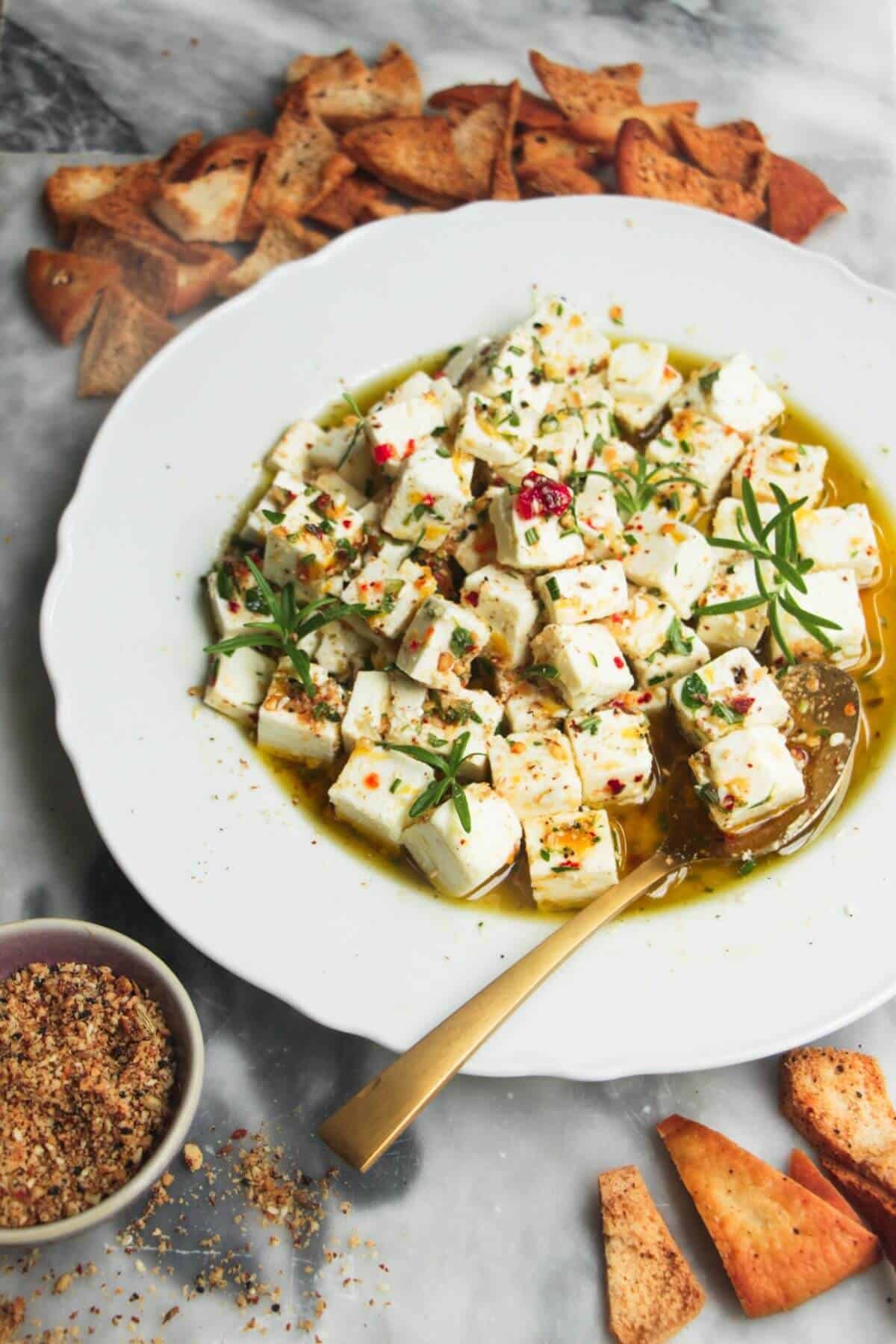 Close up of marinated feta in a white scalloped edged plate with a gold spoon inside.