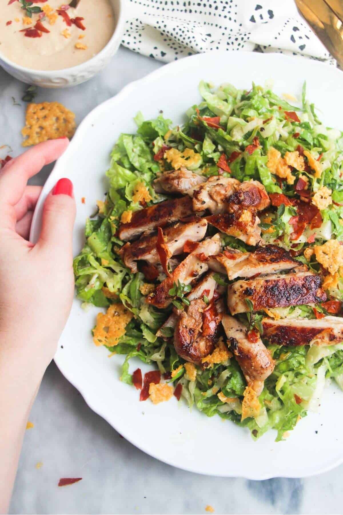 A hand holding a large white plate with sliced grilled chicken on top of green chopped lettuce.