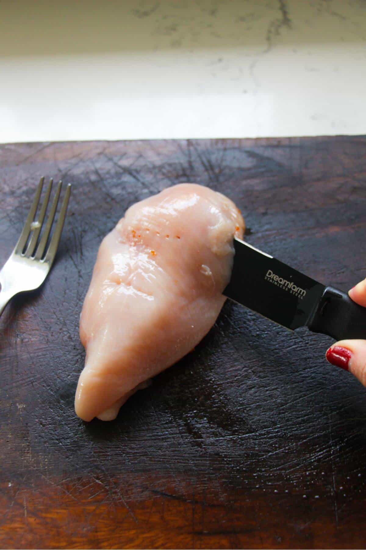 Small knife cutting a chicken breast in half on a wooden board.