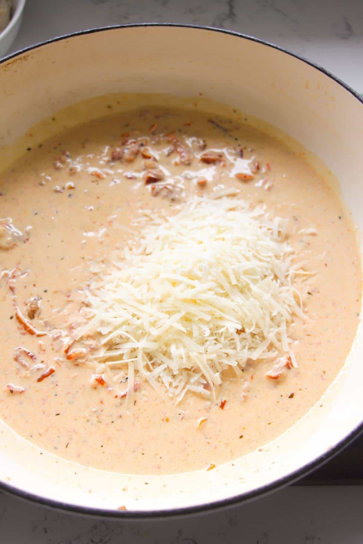 Grated parmesan cheese added to creamy sundried tomato sauce in a large white skillet.