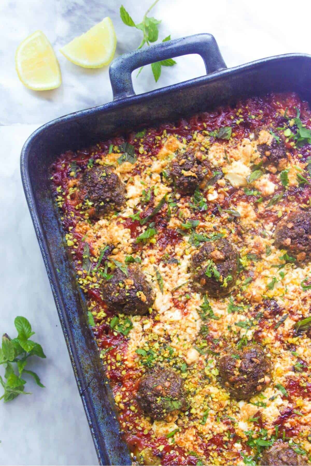 Large blue oven dish with baked meatballs in a tomato sauce, topped with feta with a lemon wedge on the side.