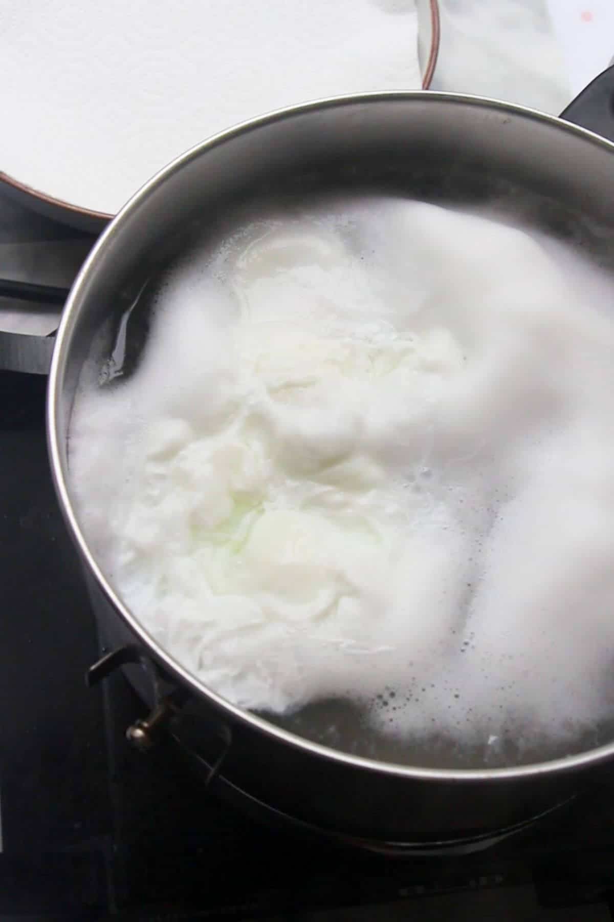 2 eggs poaching in a small silver pot with lined plate in the background.