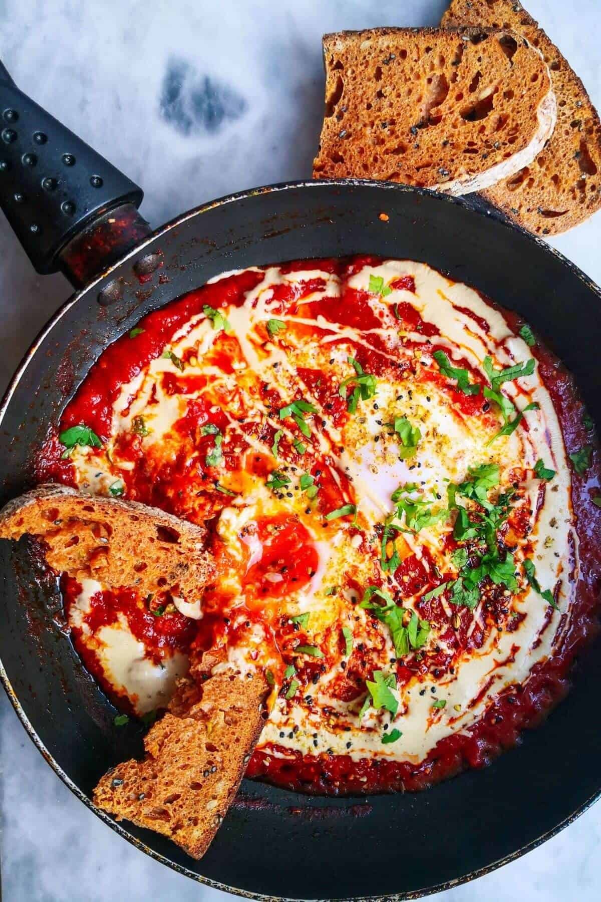 Shakshuka for one in a small black pan, on a grey marble background with sourdough placed into the sauce, and two slices of sourdough on the side.