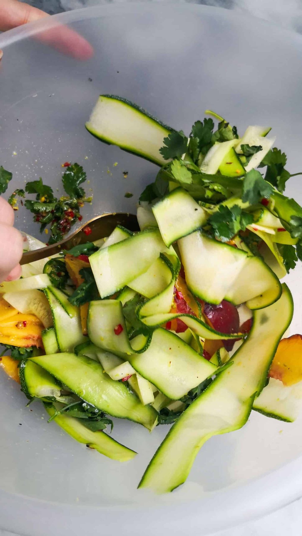 Large gold spoon mixing raw courgette and peach salad in a large clear mixing bowl.