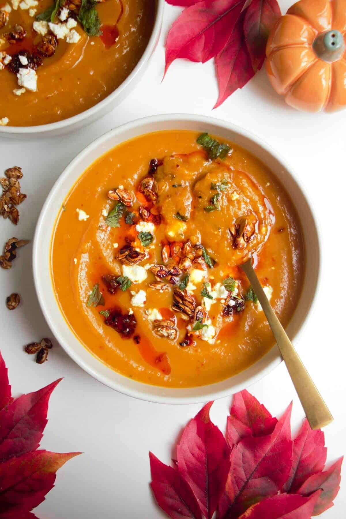 Roasted butternut squash soup in a white bowl with a gold spoon, topped with chilli oil, feta and squash seeds.