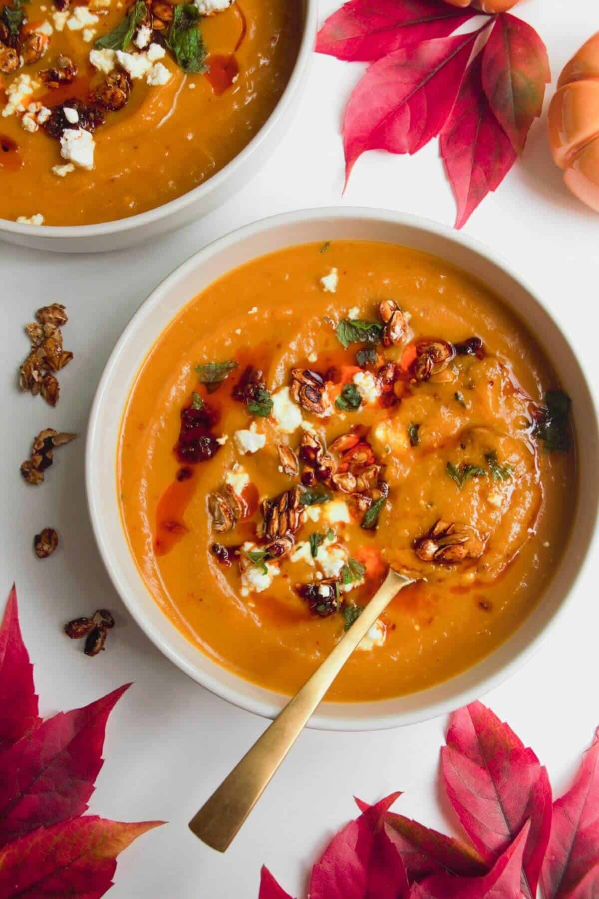 Roasted butternut squash soup in a white bowl with a gold spoon, topped with chilli oil, feta and squash seeds.