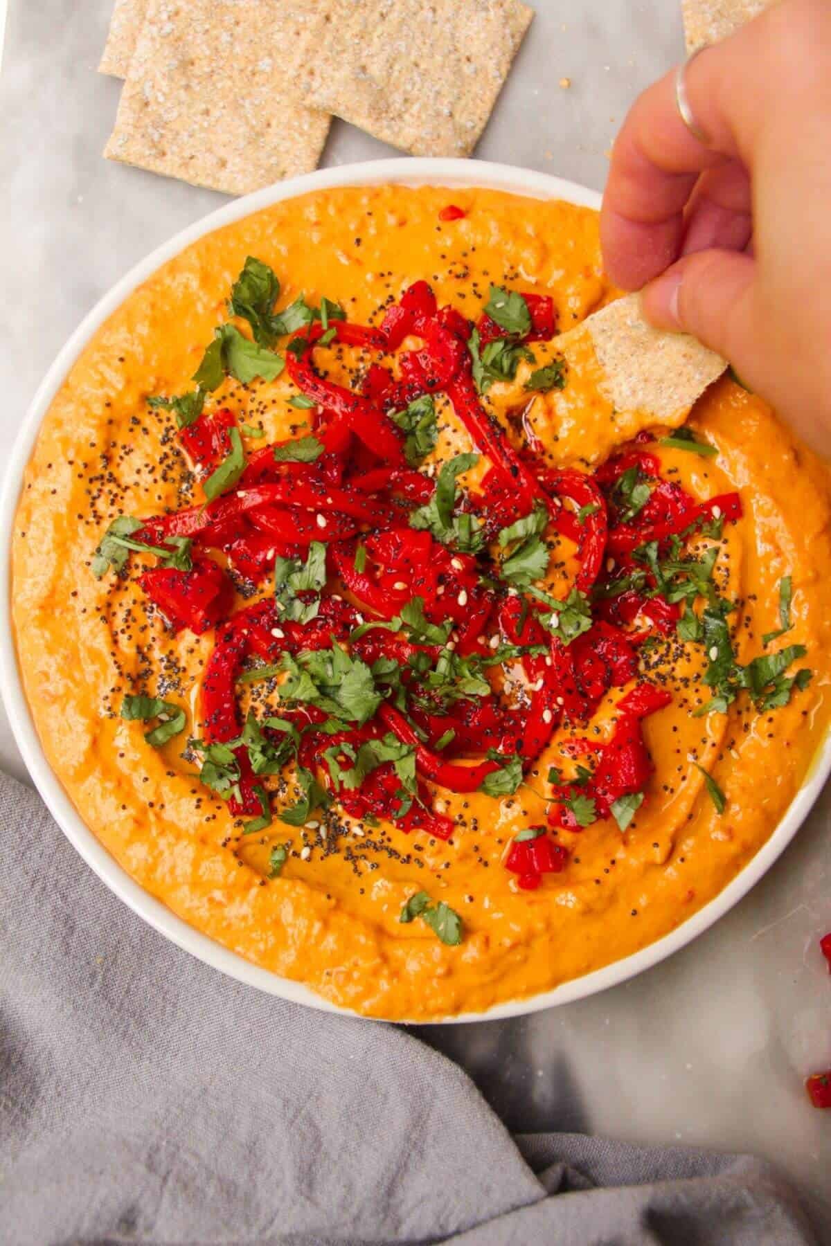 Hand scooping cracker in red pepper hummus on a white plate.