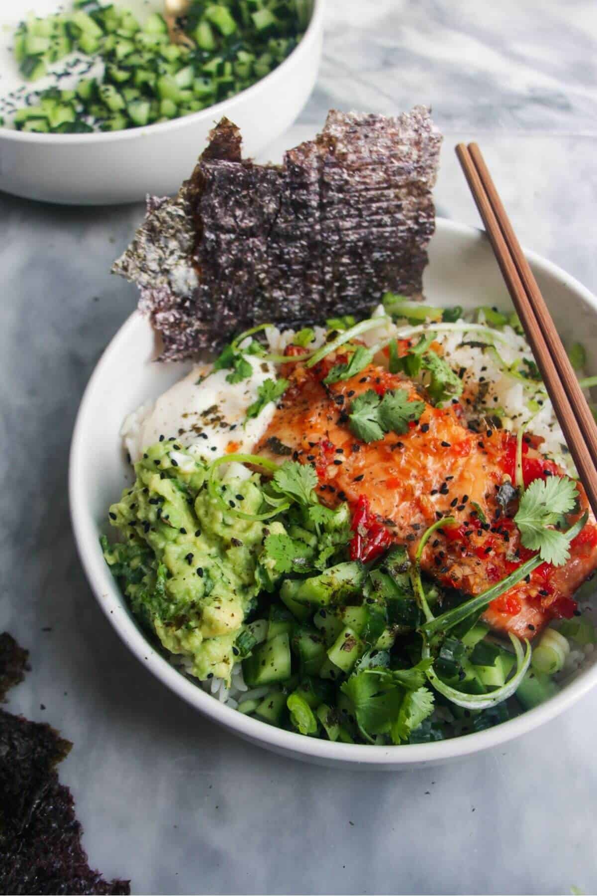 White bowl filled with colourful vegetables and a salmon fillet with seaweed sheets on the side and chopsticks in the bowl.