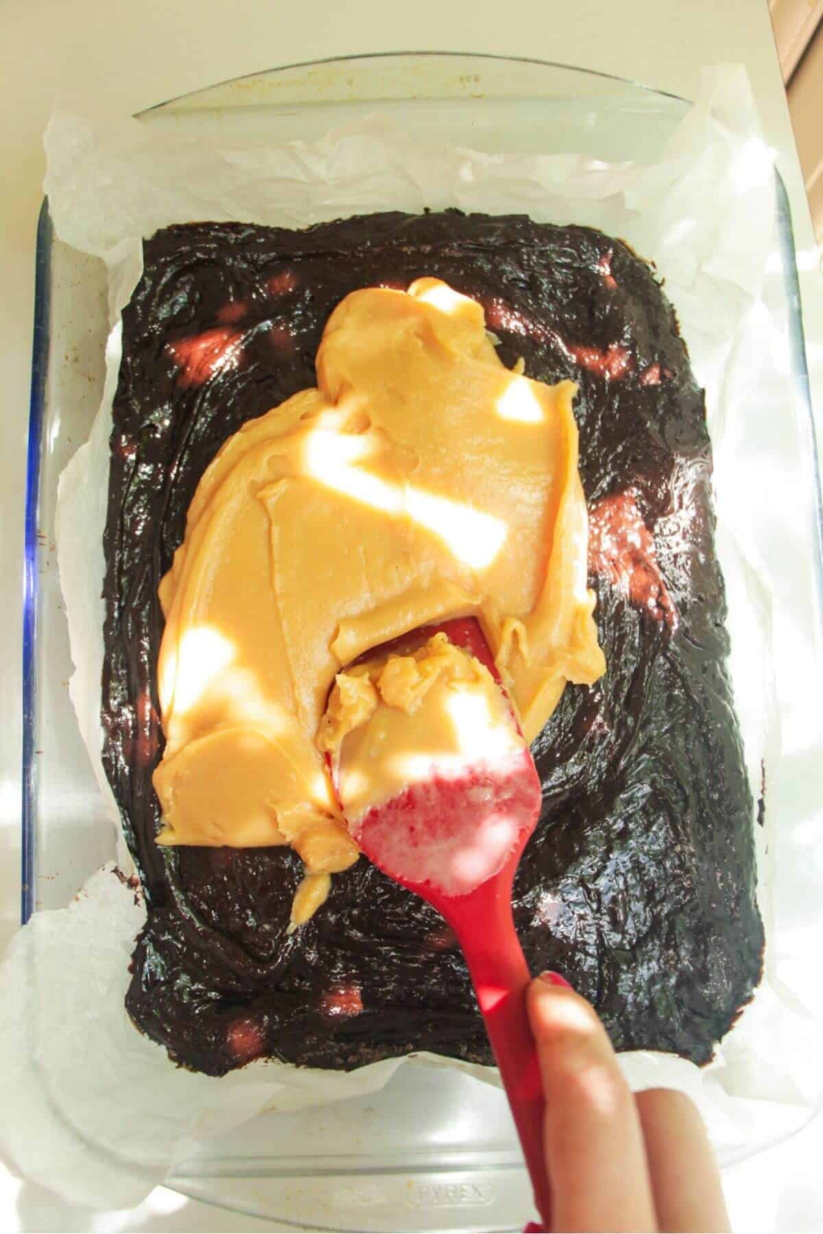 Spatula spreading salted caramel on top of brownie batter in an oven dish.