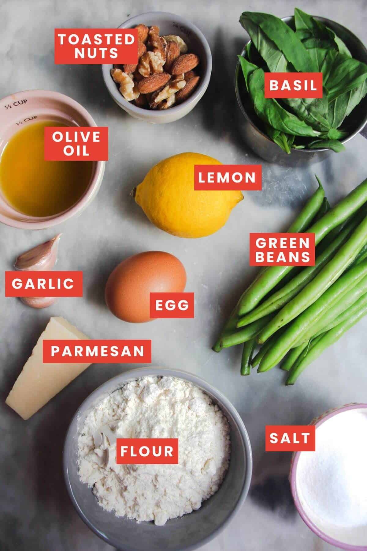 Ingredients for scissor cut pesto pasta on a marble background and labelled.