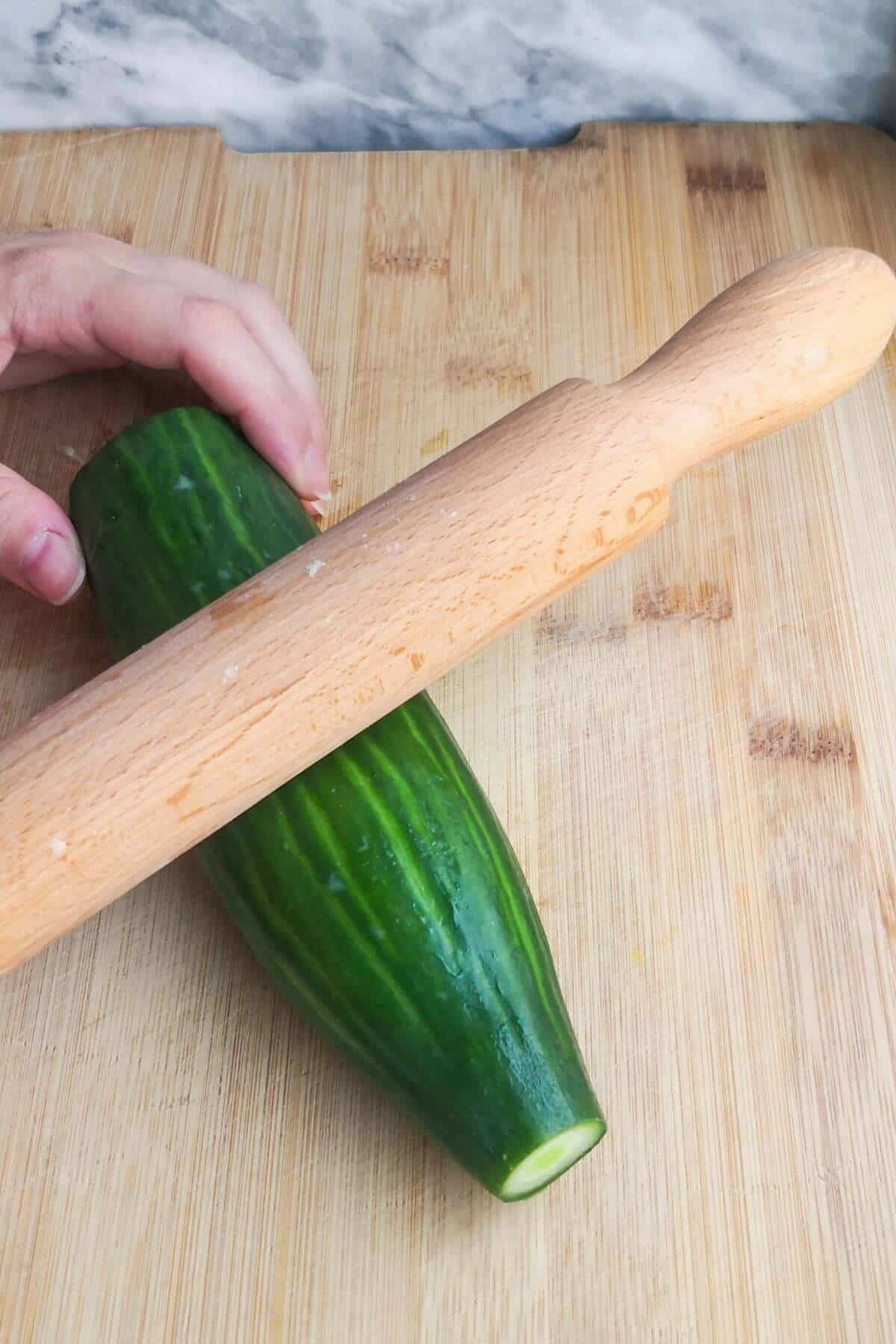 A hand holding half a cucumber and a wooden rolling pin pressing down on it.
