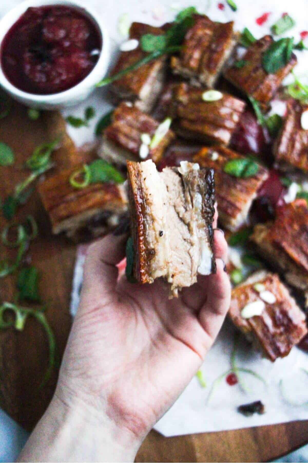 Hand holding up a piece of crispy slow roasted pork belly, showing a cross-section of the meat.
