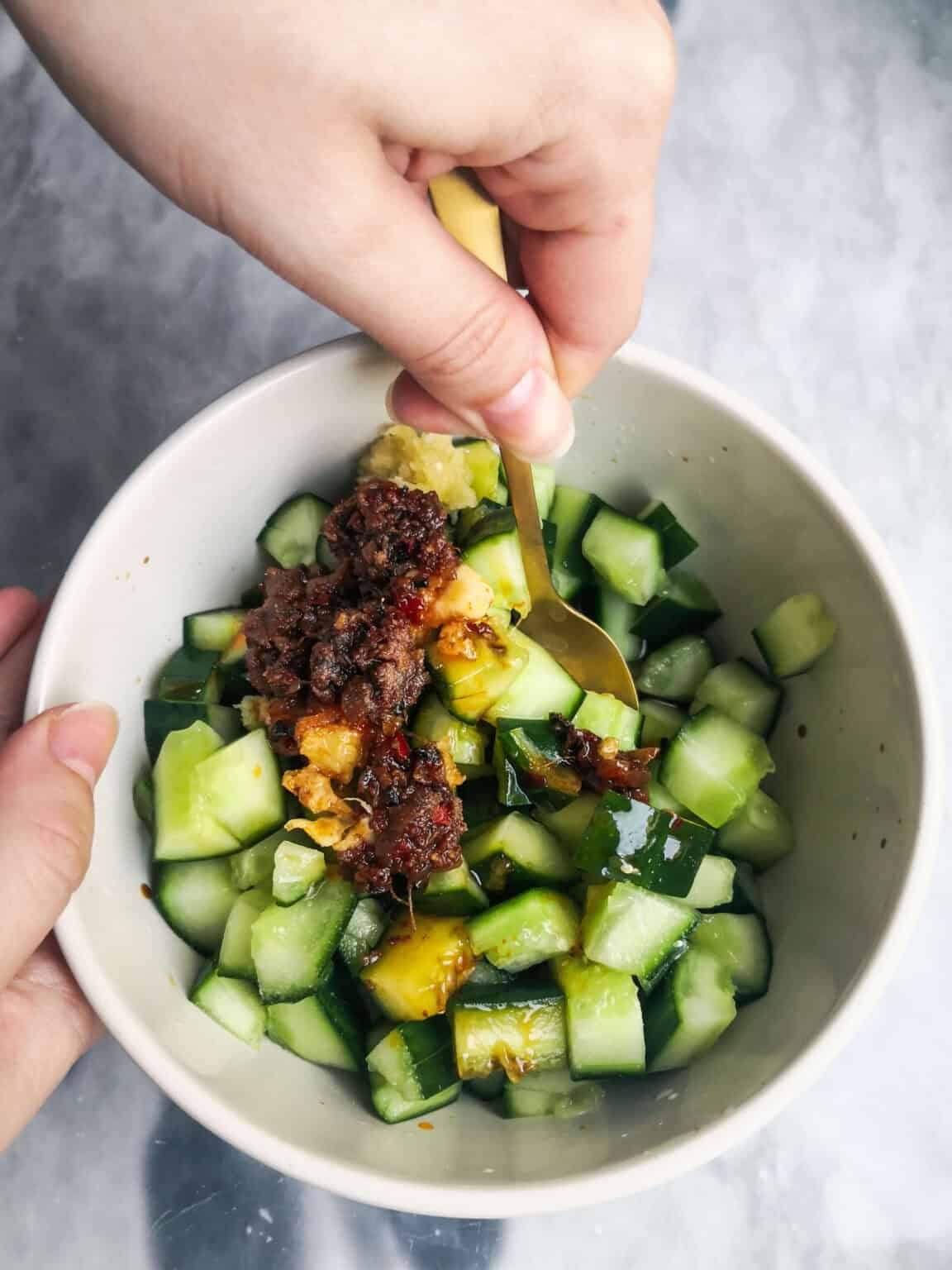 Hand holding a small gold spoon mixing the cucumber salad in a small white bowl.