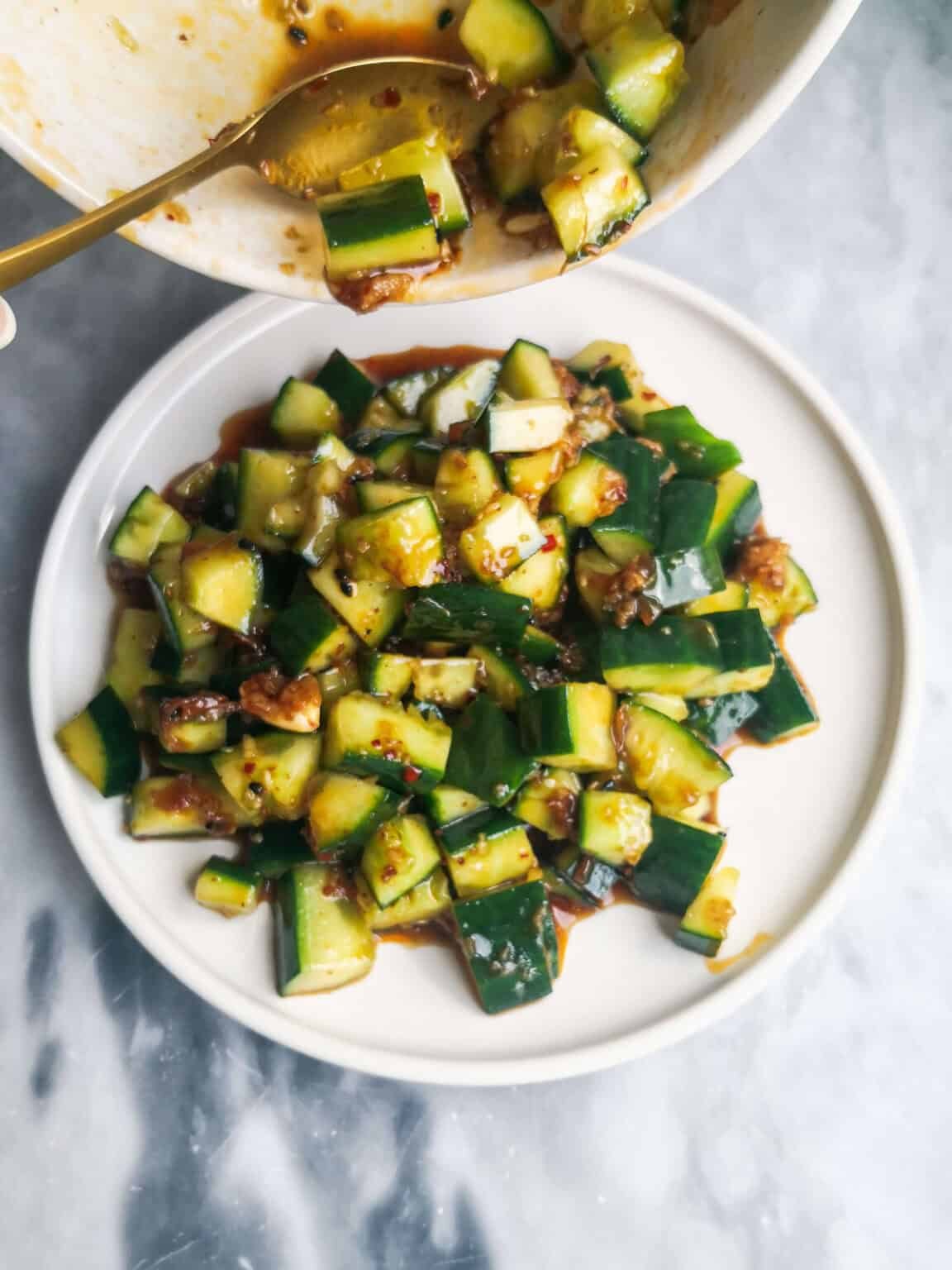 Gold spoon pouring the smacked cucumber salad on a small white serving plate on a grey marble background.