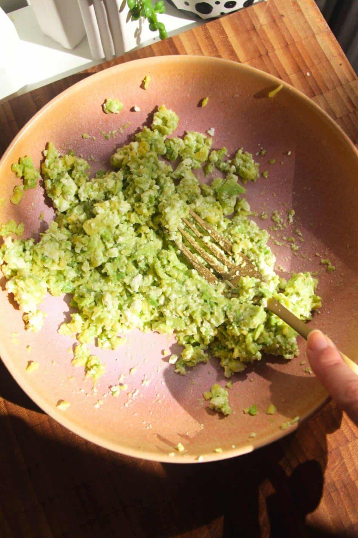 Fork mashing avocado and feta in a pink bowl.