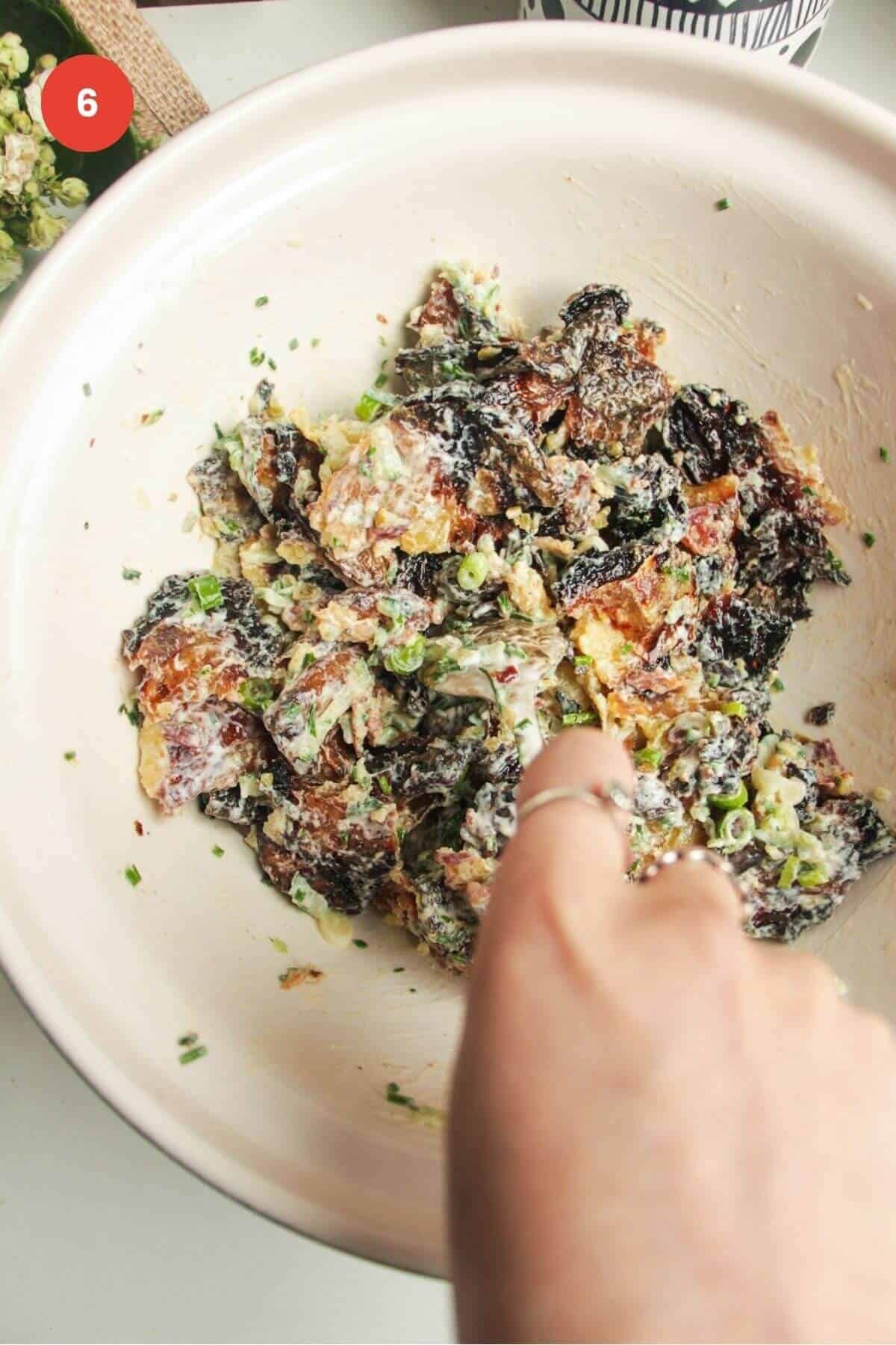 Hand holding spoon mixing smashed potato salad in a bowl.