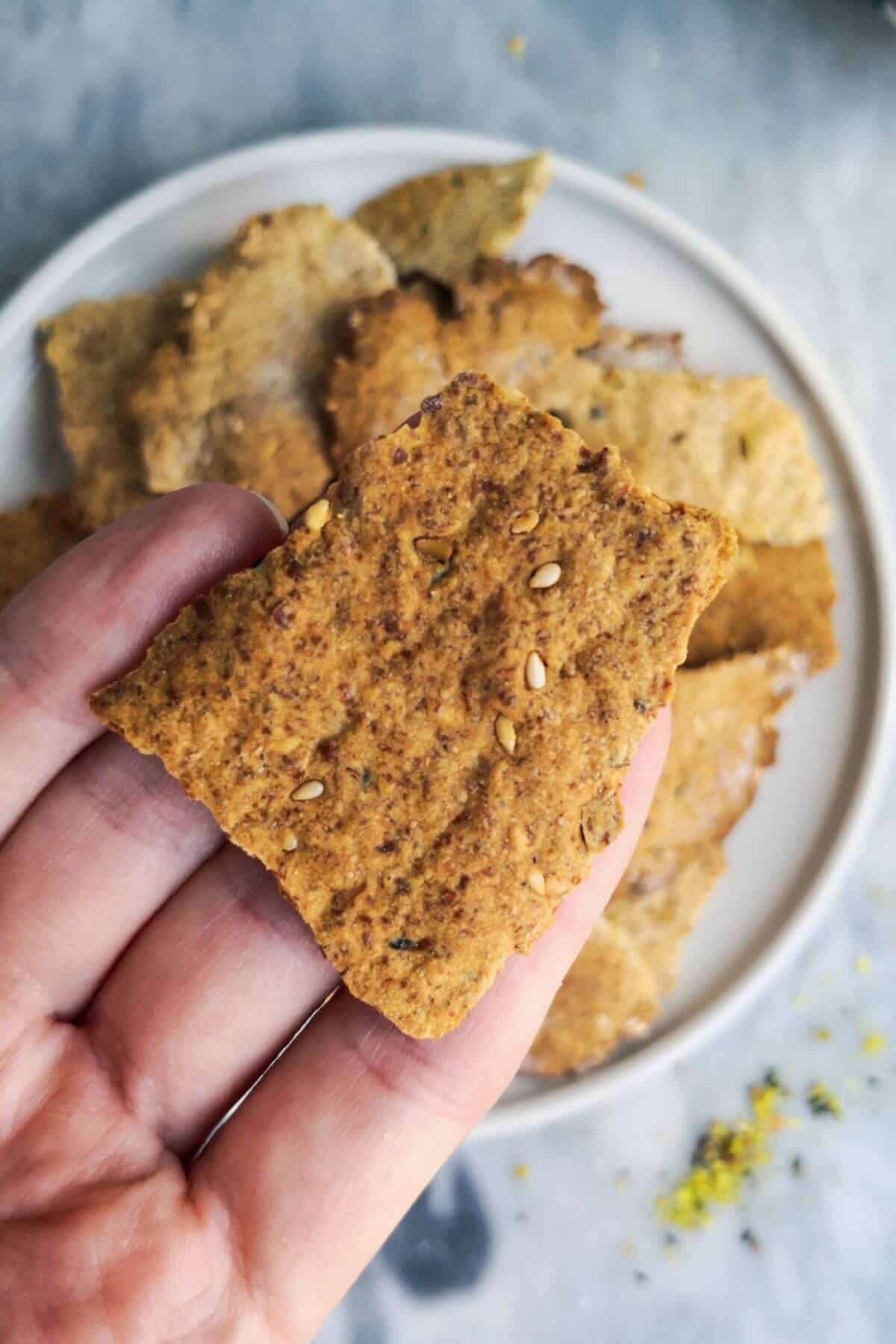 Close up of sourdough cracker in a hand with a plate of crackers in the background.