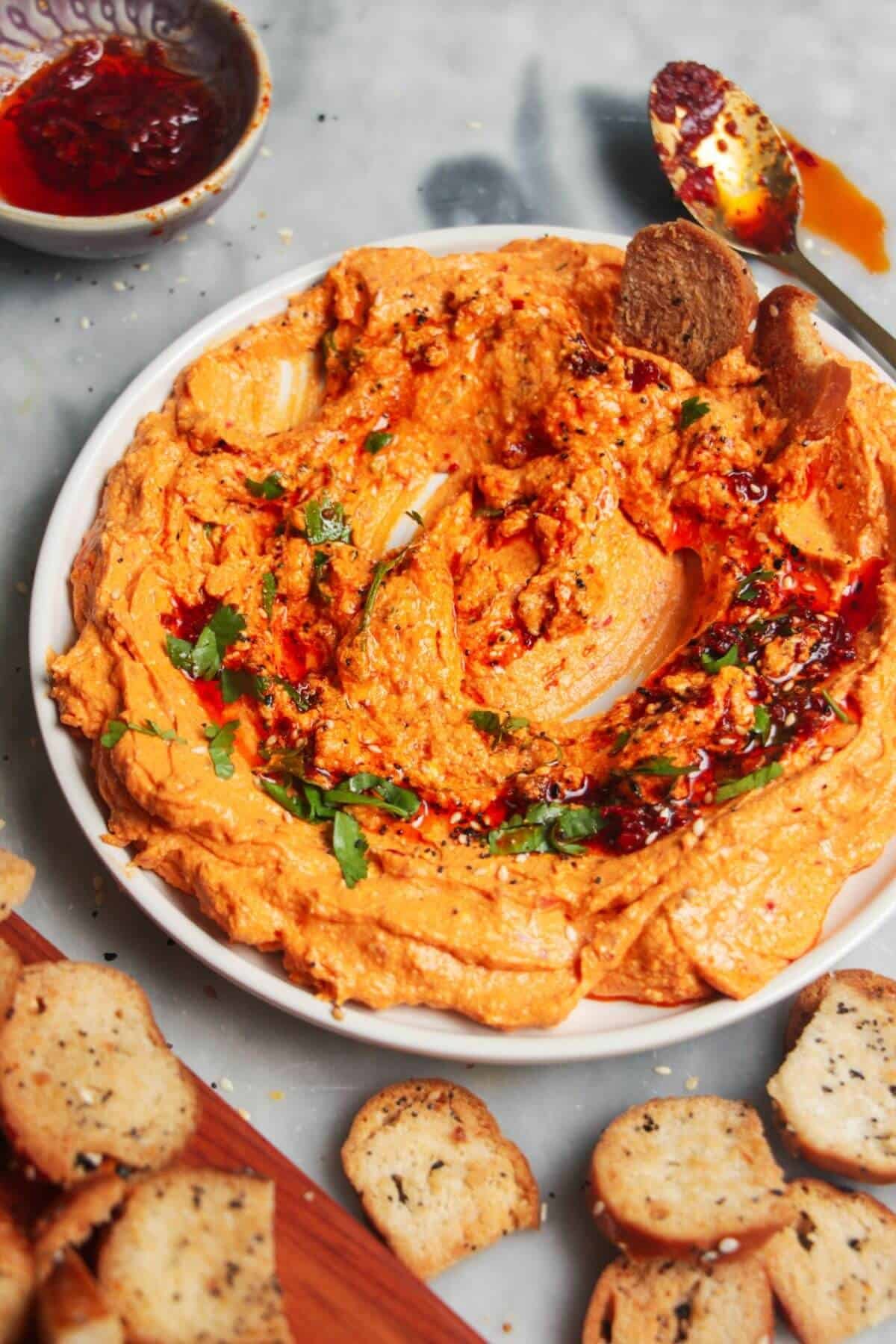 Spicy harissa feta dip on a small white plate, with bagel chips and more harissa on the side.