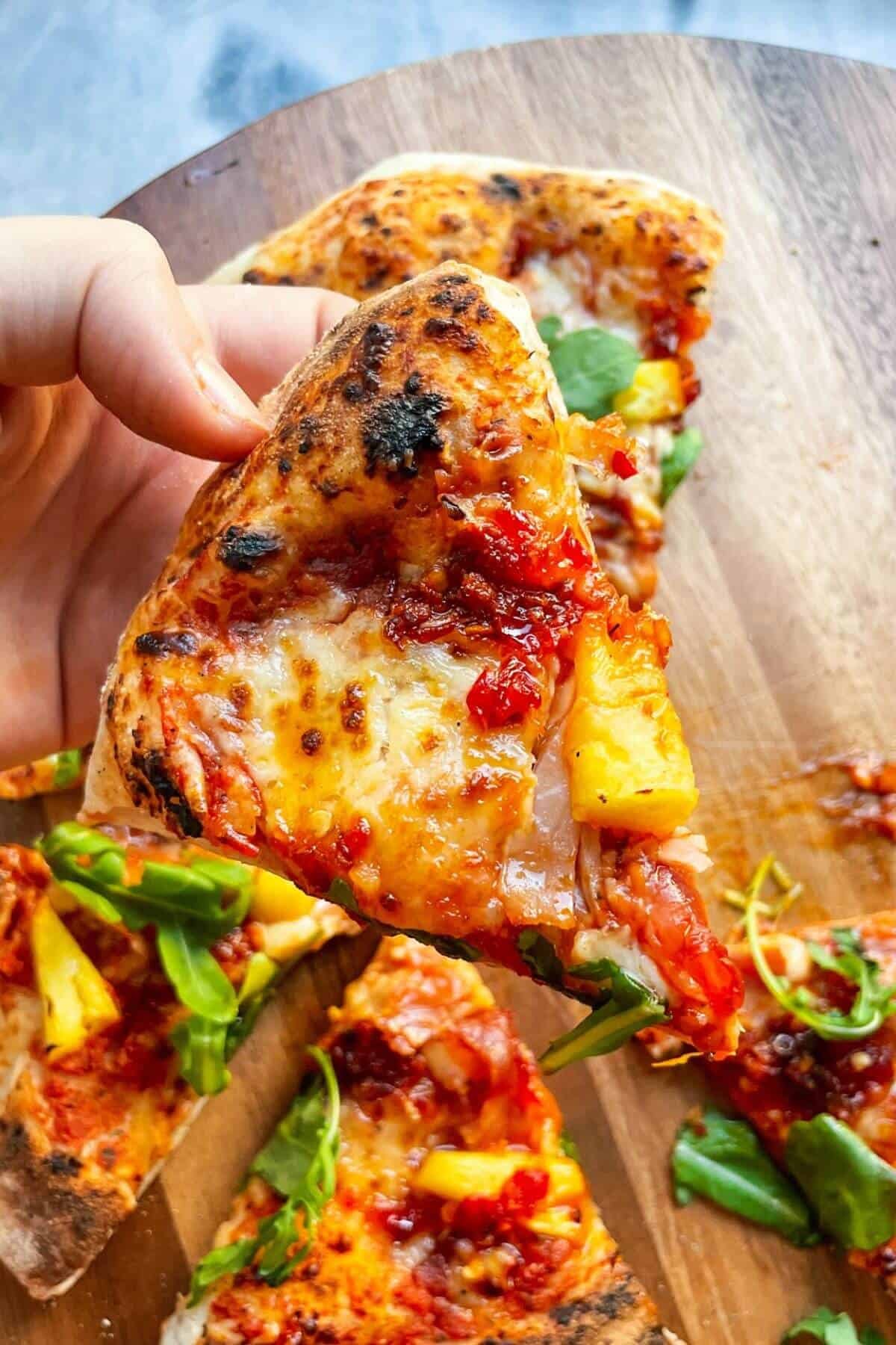 Hand holding up a piece of spicy Hawaiian pizza with pizza in the background.