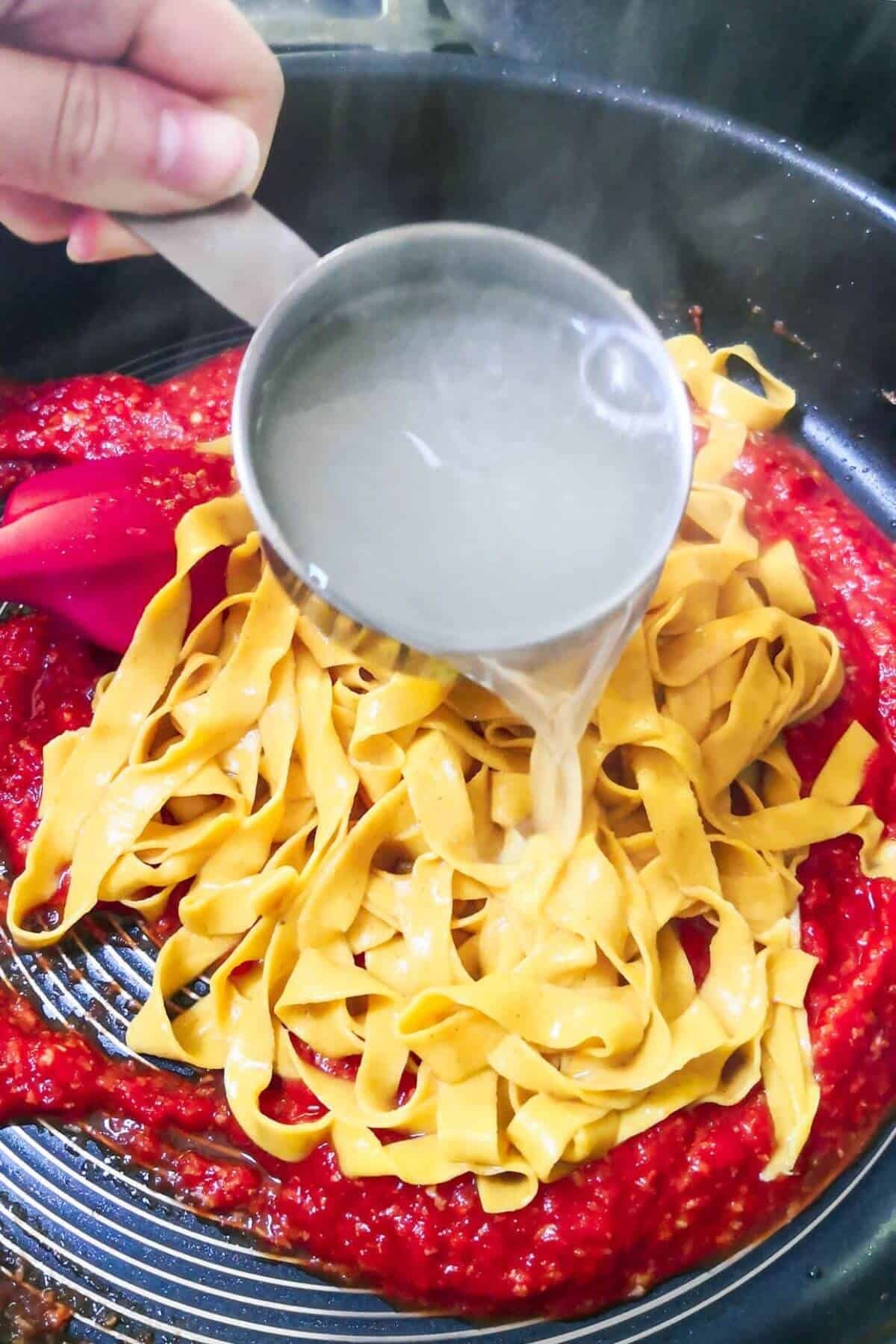 A small cup of pasta water being added to pasta and nduja pasta sauce in a large pan.