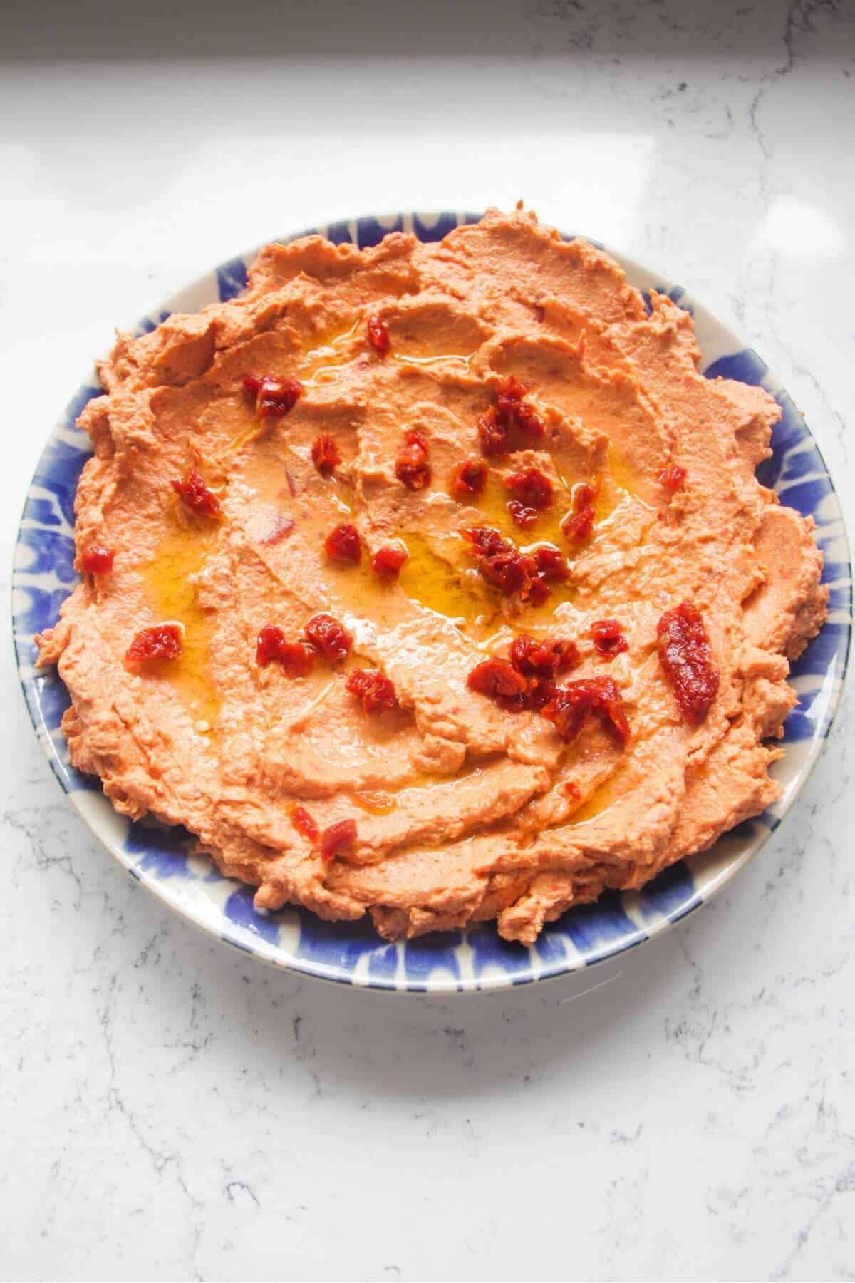 Chopped sundried tomatoes on top of sundried tomato feta dip smoothed out onto a small blue and white plate.