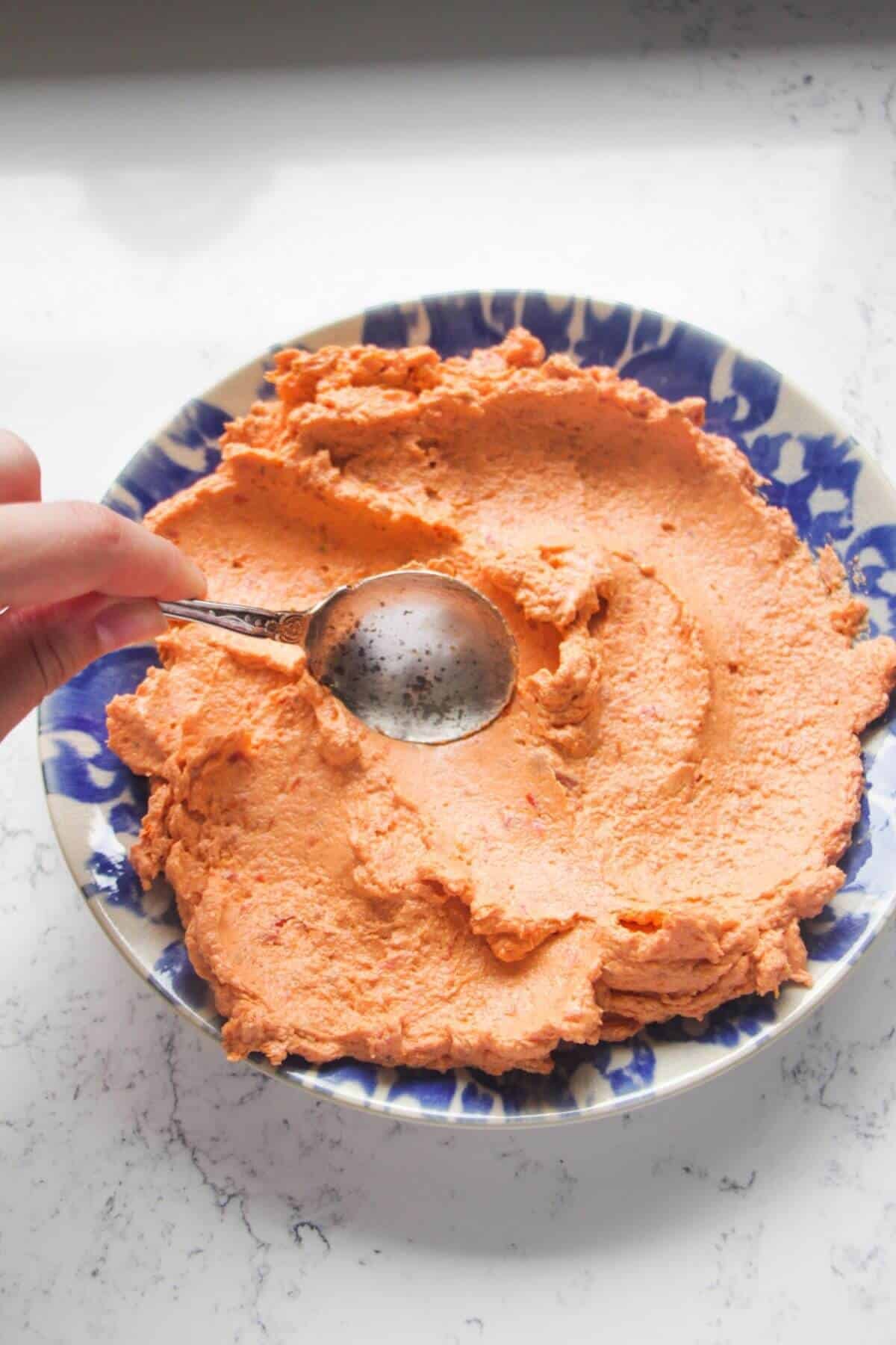 Silver spoon smooth sundried tomato feta dip onto a small white and blue plate.