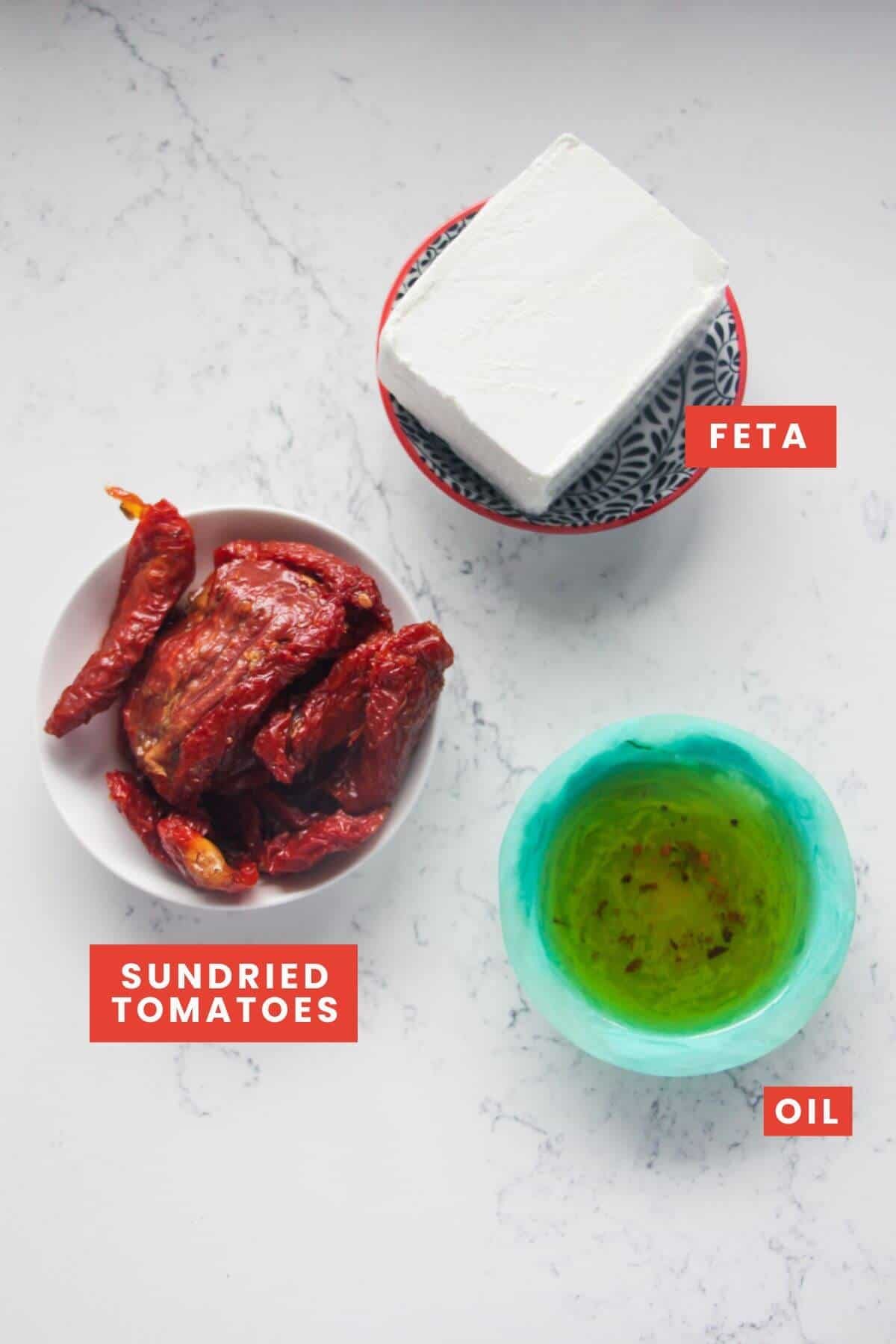 Ingredients for sundried tomato feta dip laid out on white marble background and labelled.