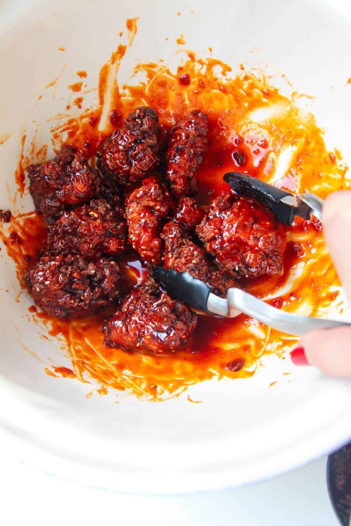 Tongs mixing fried chicken with sticky glaze in a large white bowl.
