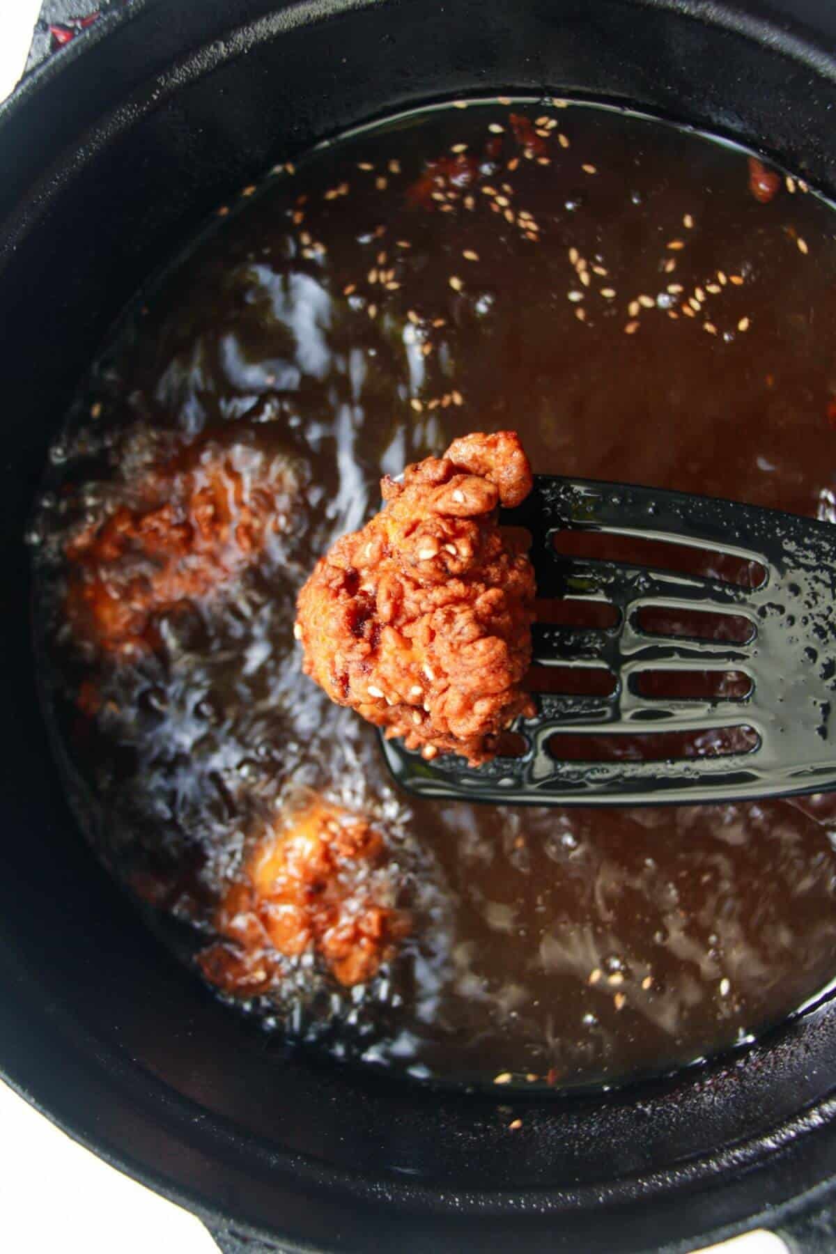Spatula holding up a piece of golden fried chicken with more chicken in the background.