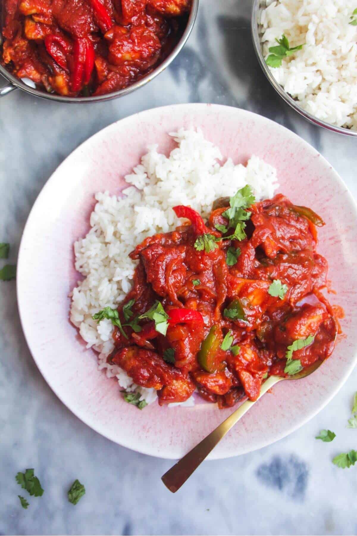Chicken jalfrezi curry on top of rice on a small pink plate, with a gold spoon.