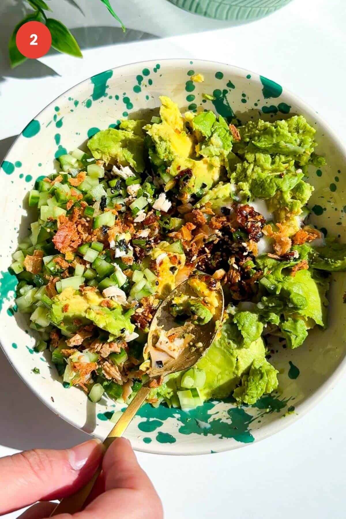 Avocado, shredded chicken, chili crisp, sesame seeds, cucumber being mixed in a bowl.
