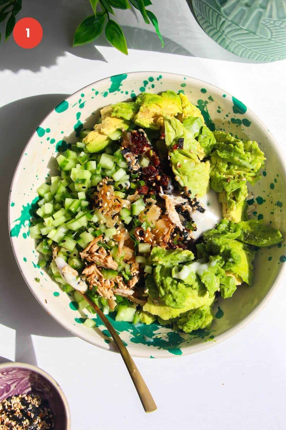Avocado, shredded chicken, chili crisp, sesame seeds, cucumber in a bowl.