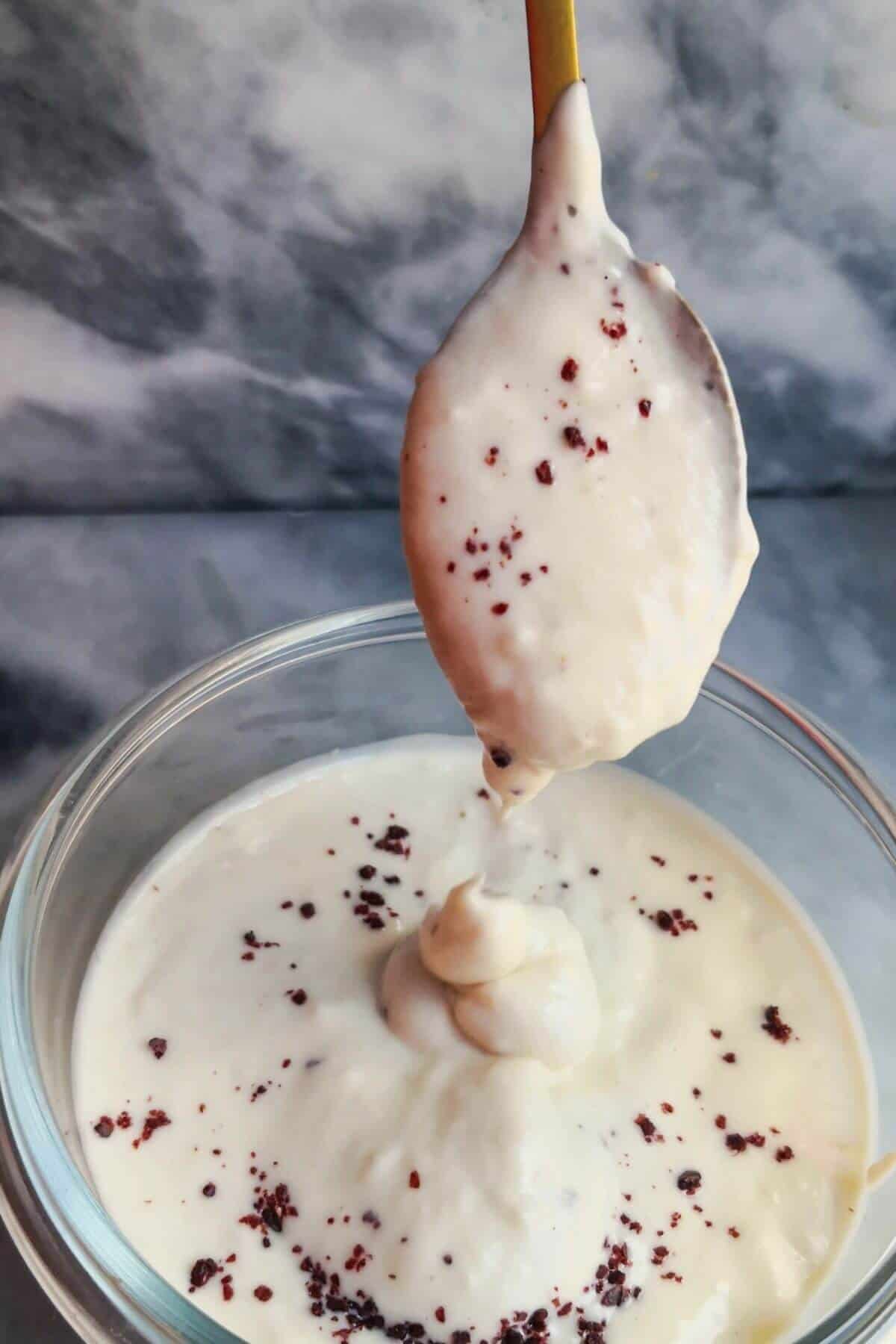 Tahini yogurt sauce being poured off a small gold spoon into a small glass bowl on a grey marble background.