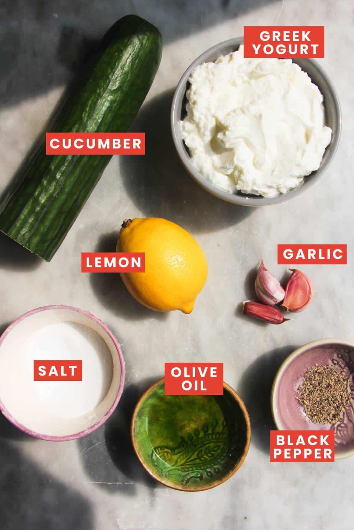 Ingredients for tzatziki sauce laid out on a grey marble background and labelled.