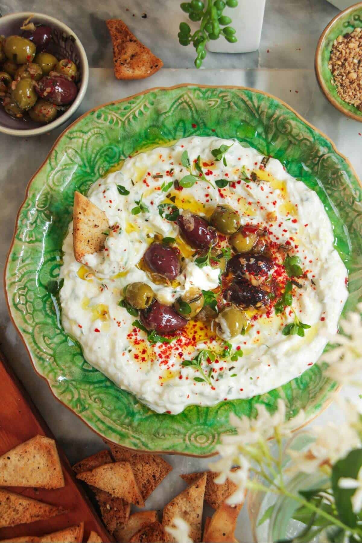 Tzatziki sauce spread onto a small green serving plate, topped with marinated olives with pita chips on the side.