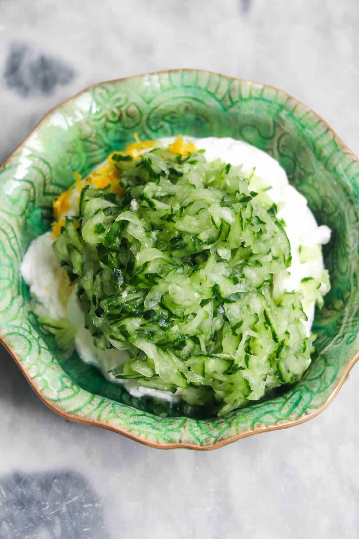 Grated cucumber, yogurt and lemon zest in a small green bowl on a grey marble background.