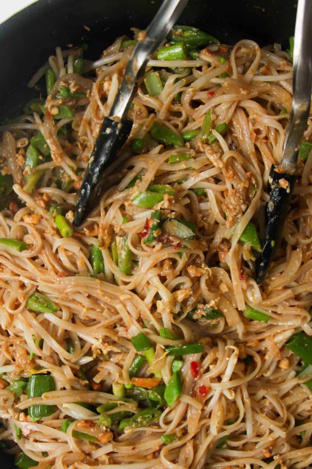 Tongs tossing scallions and peanuts through Pad Thai in a large frying pan.