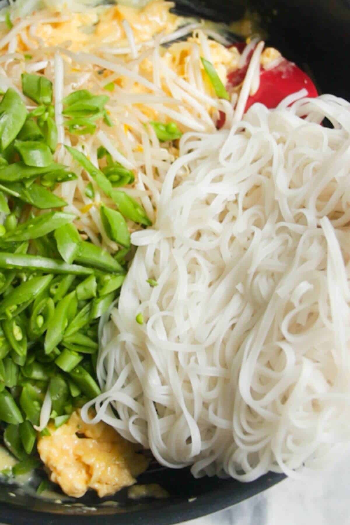 Rice noodles, sugar snap peas added to scrambled eggs and bean sprouts in a large frying pan.
