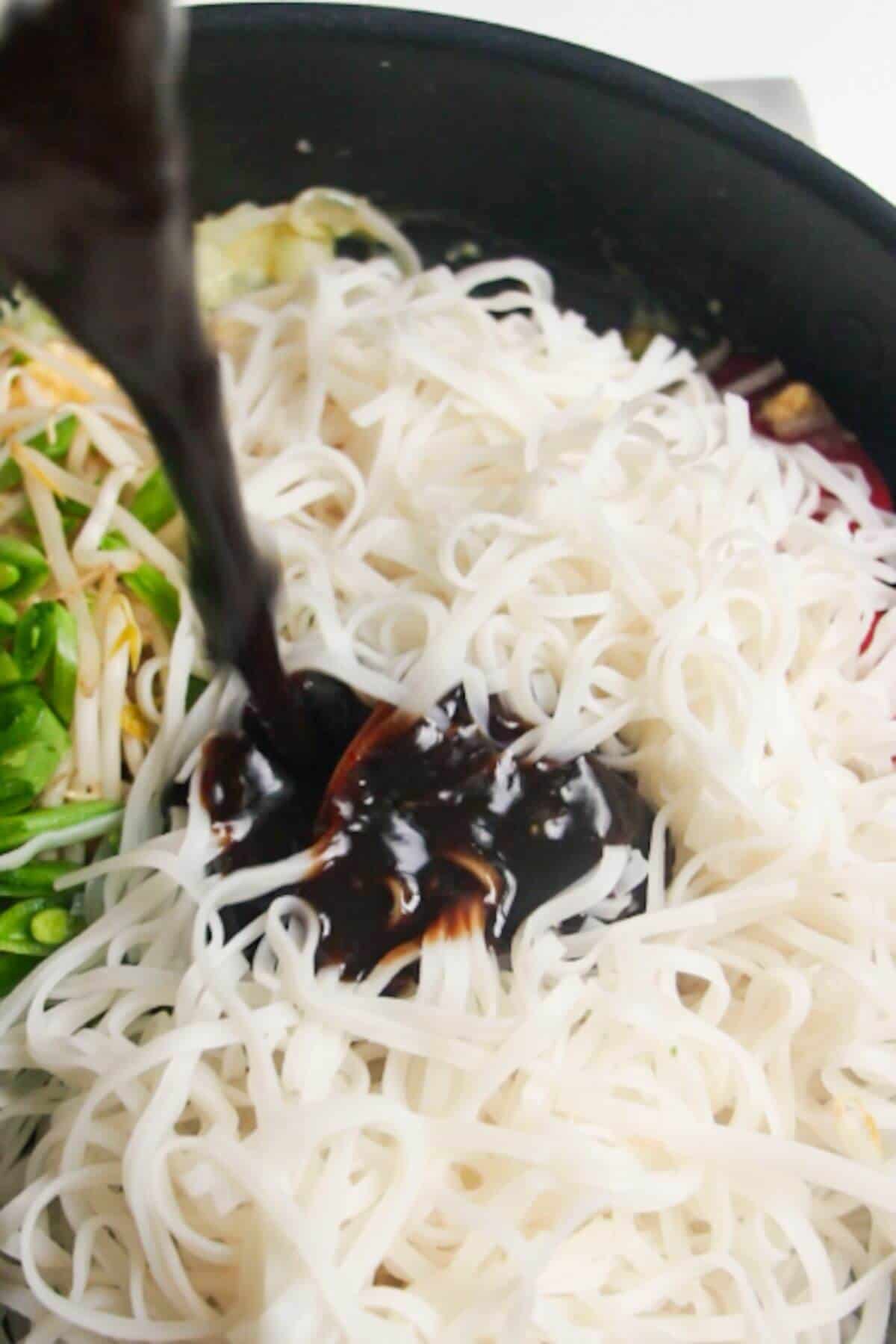 Dark brown sauce being poured on top of rice noodles in a large frying pan.