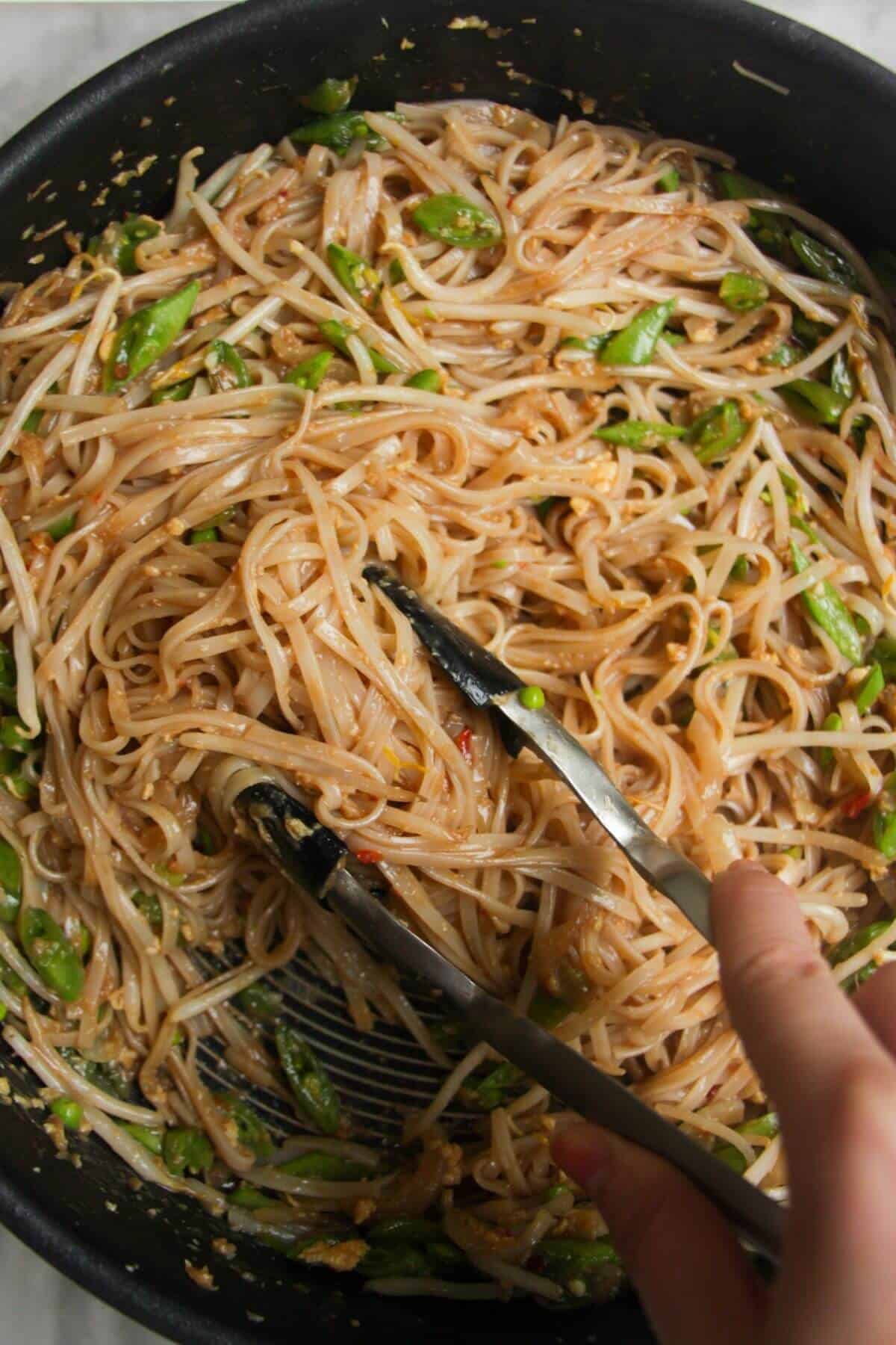 Tongs tossing noodles and veggies in a large frying pan.