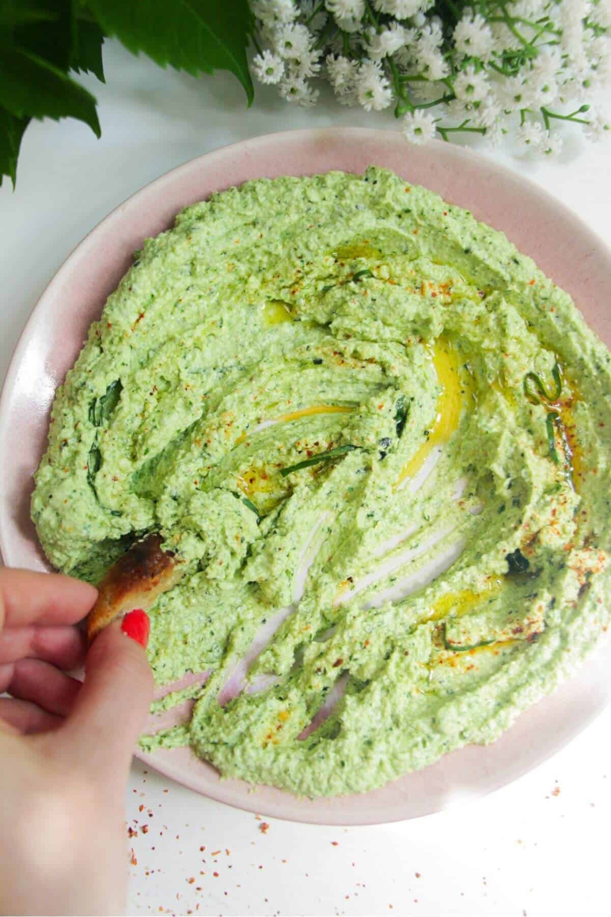 Whipped feta pesto dip on a pink plate with a small piece of bread on thes side.
